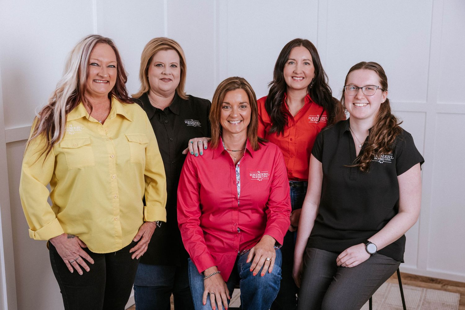 Five professional women posing together for a group photo, wearing colorful shirts in a bright, modern interior setting.