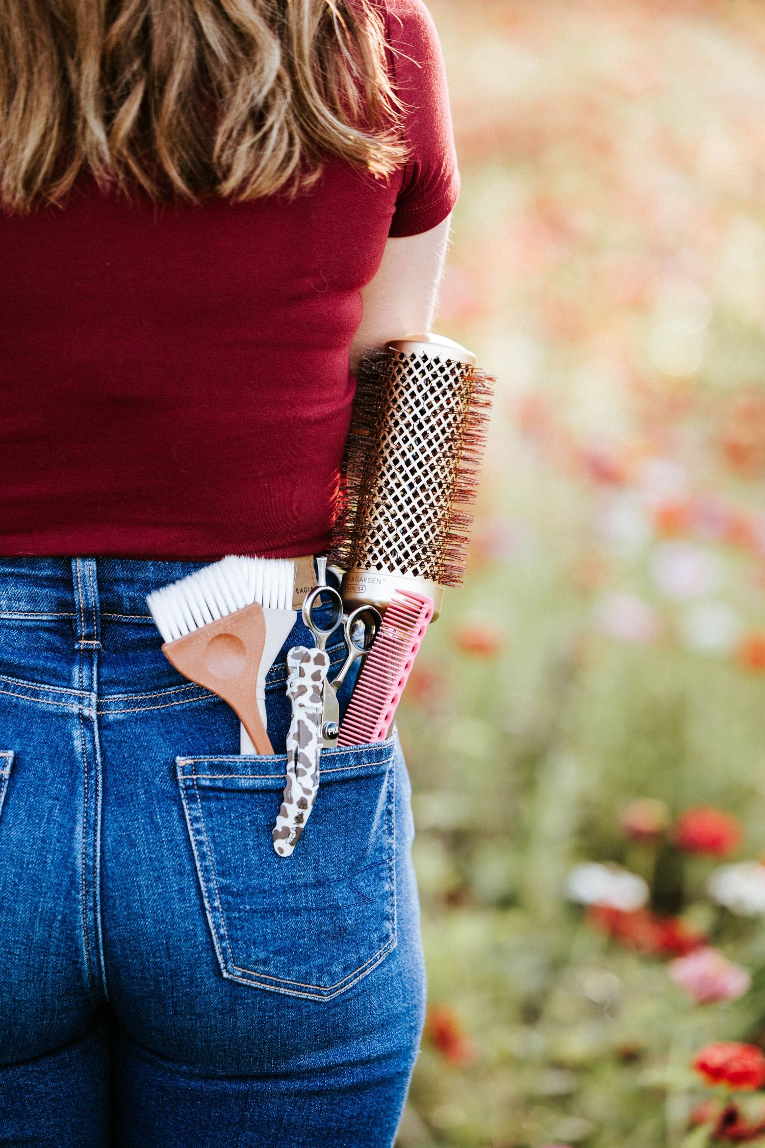 Woman in jeans holding hairstyling tools including brushes and scissors in back pocket outdoors.