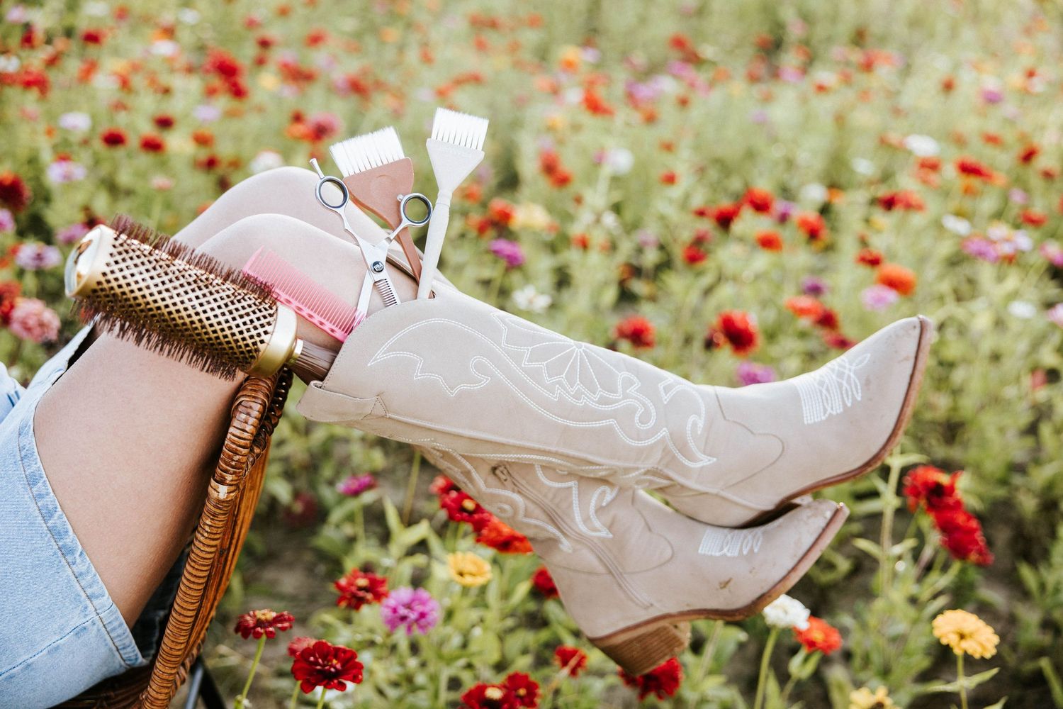 White cowboy boots with embroidery resting on a knee in a vibrant flower field with red, pink, and yellow blooms.