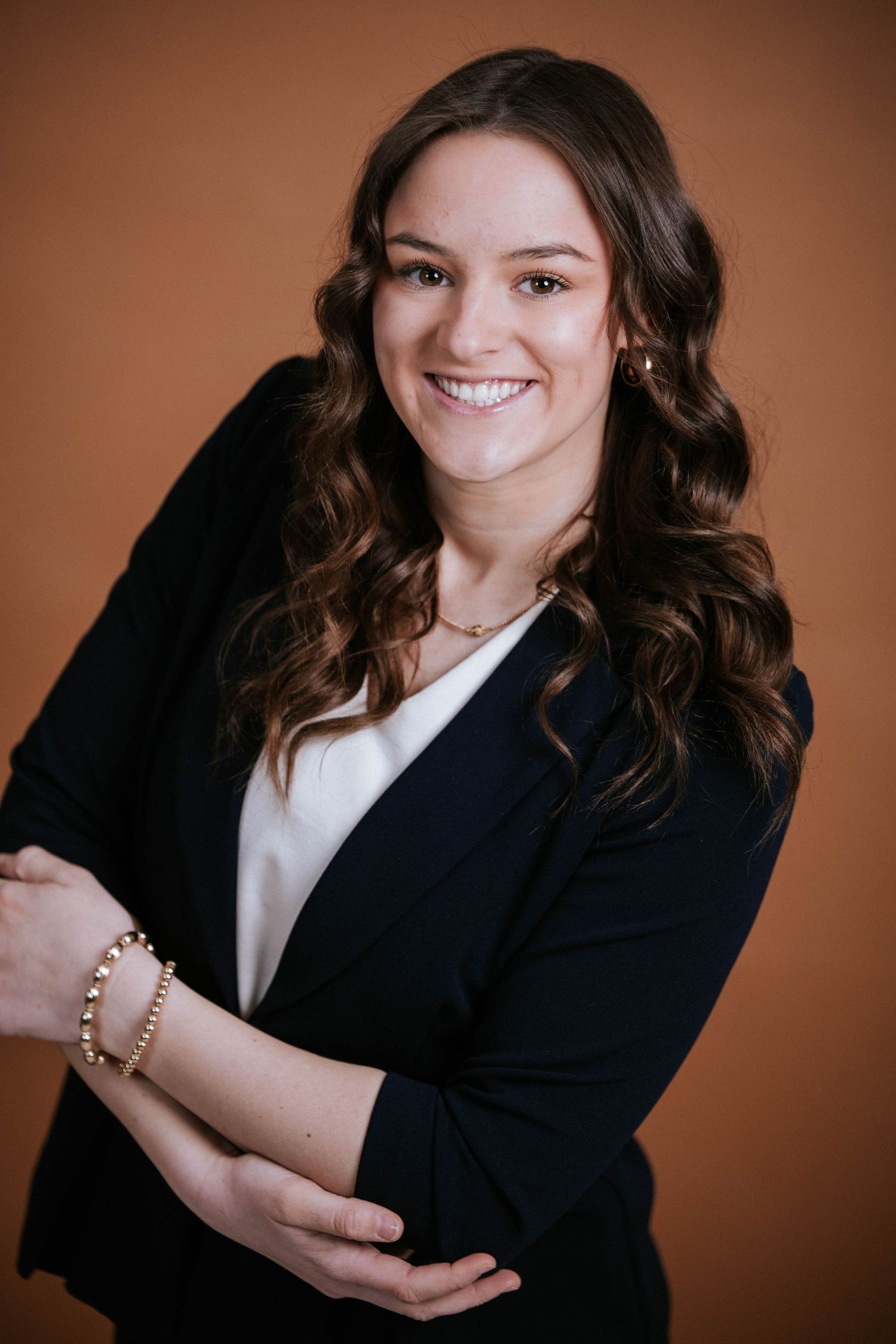 Professional woman in black blazer smiling confidently against warm brown background, wearing gold jewelry.