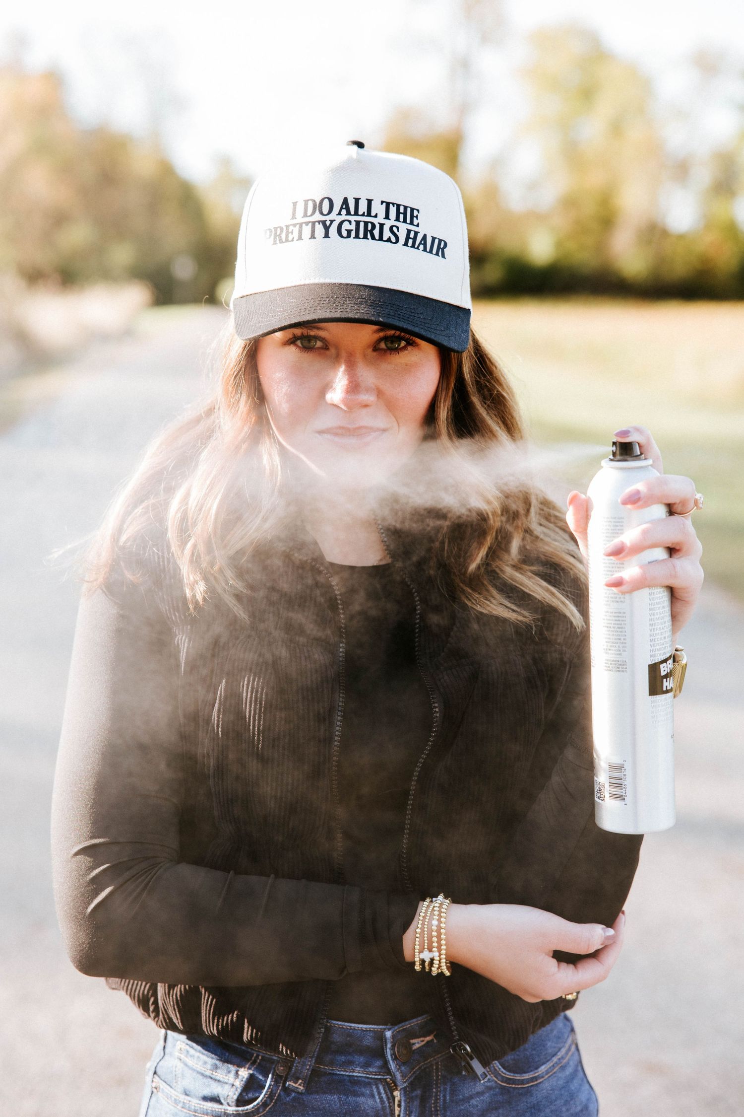 Woman in 'I Do All The Pretty Girls Hair' cap spraying hairspray outdoors.