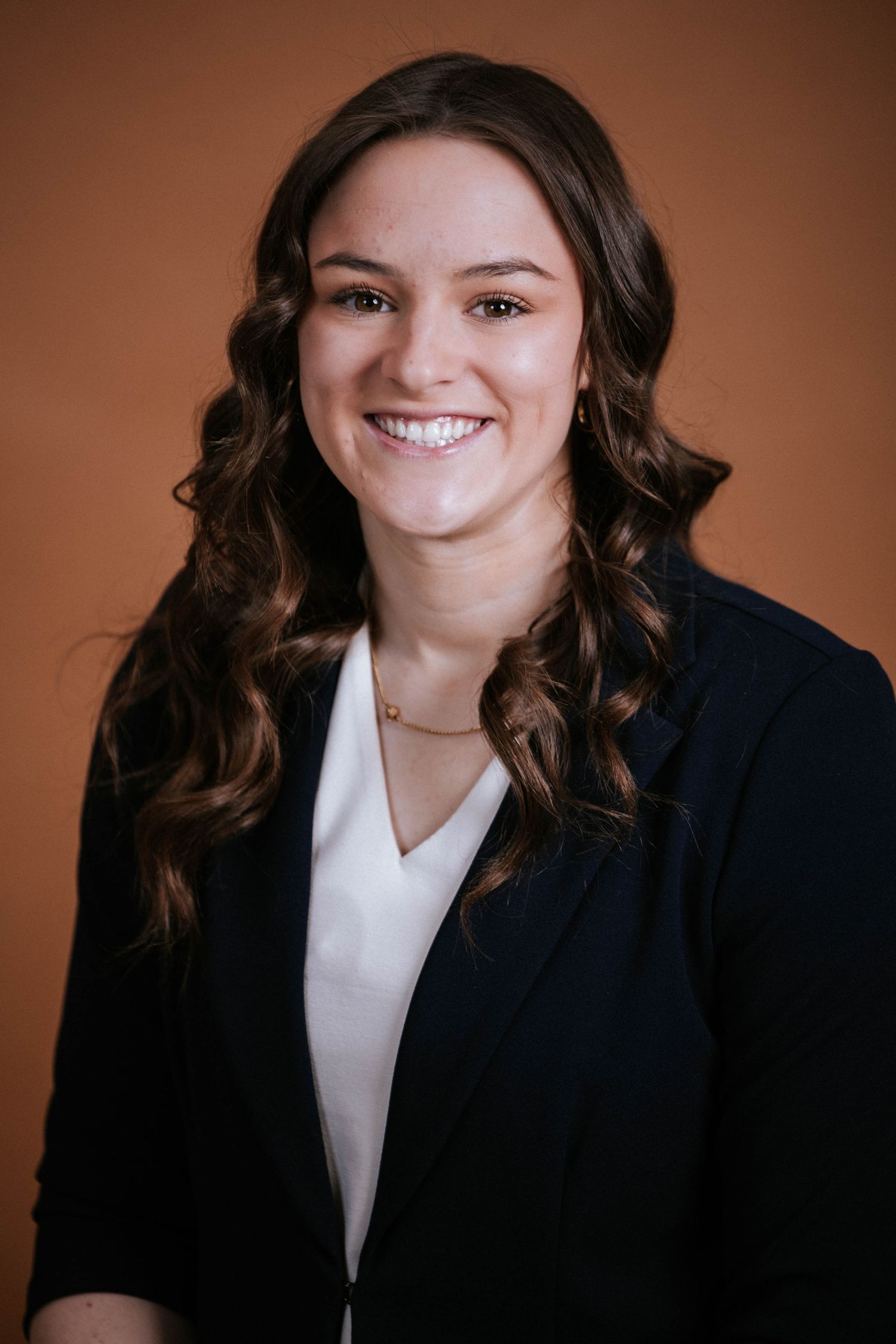 Professional headshot of a smiling young woman with curly brown hair wearing a navy blazer and white blouse.