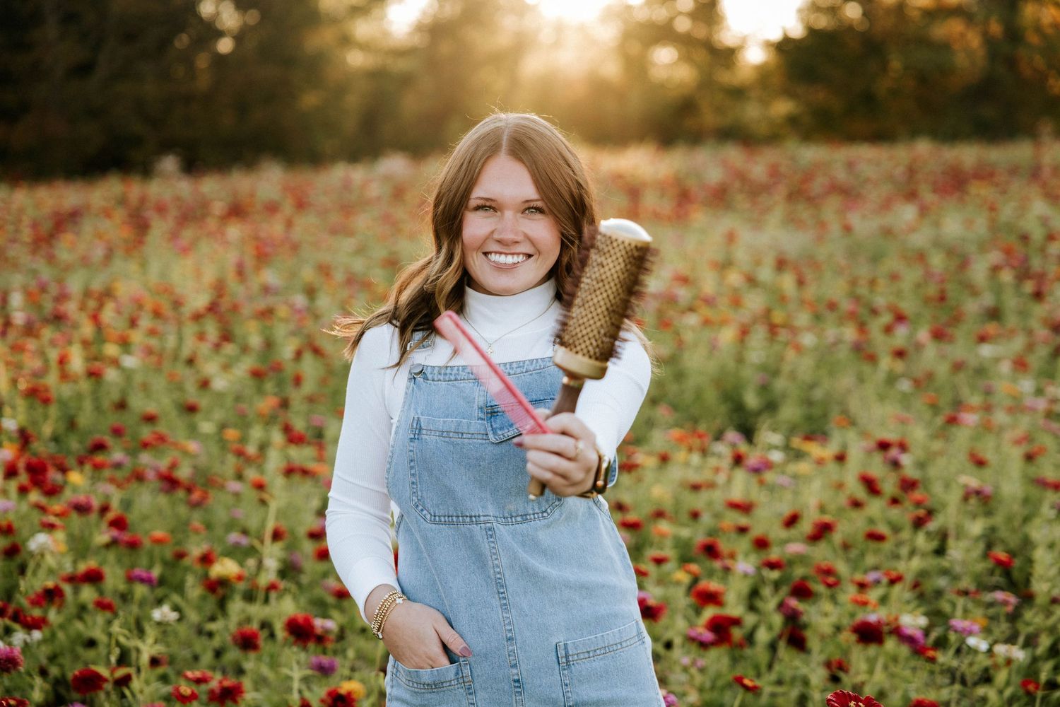 Smiling young woman in denim overalls holding a brush in a vibrant flower field during golden hour.