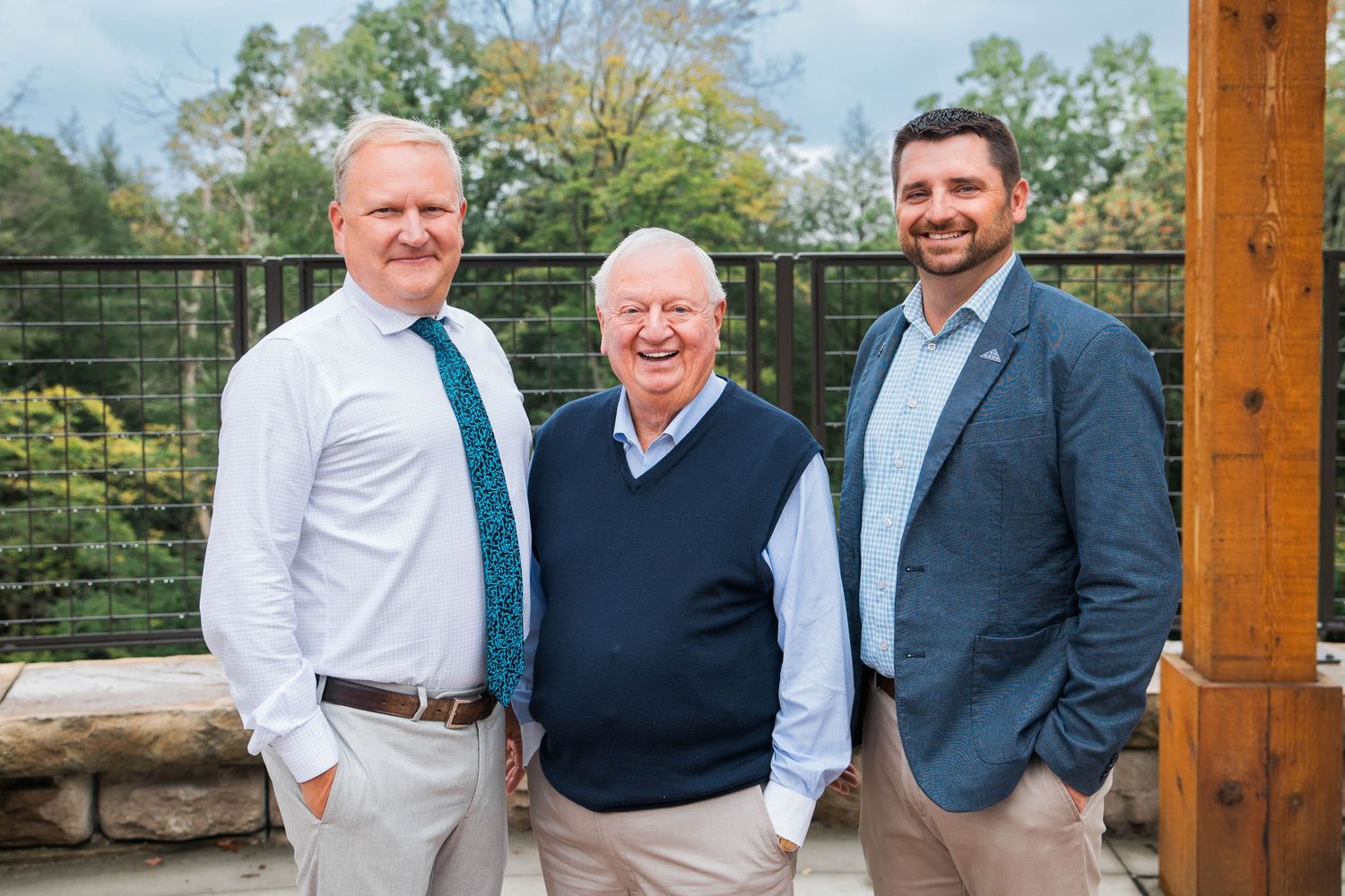 Three smiling men in business casual attire pose together outdoors on a stone patio surrounded by lush greenery.