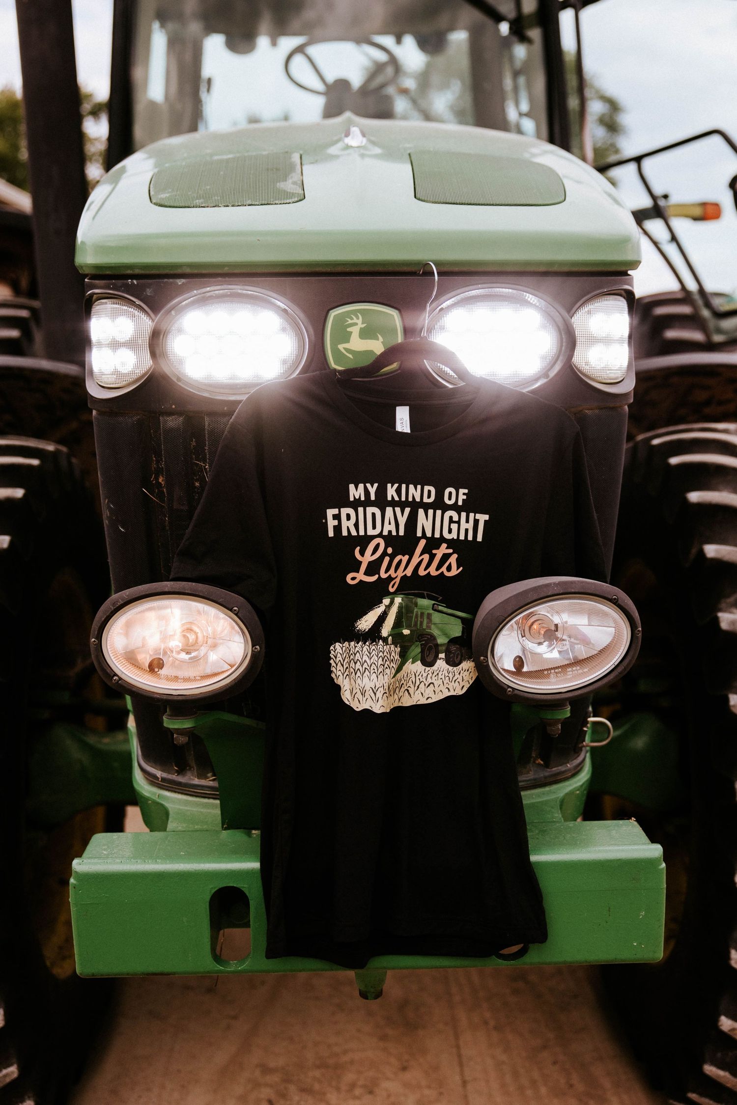 John Deere tractor with lit headlights displaying a black t-shirt reading My Kind of Friday Night Lights.