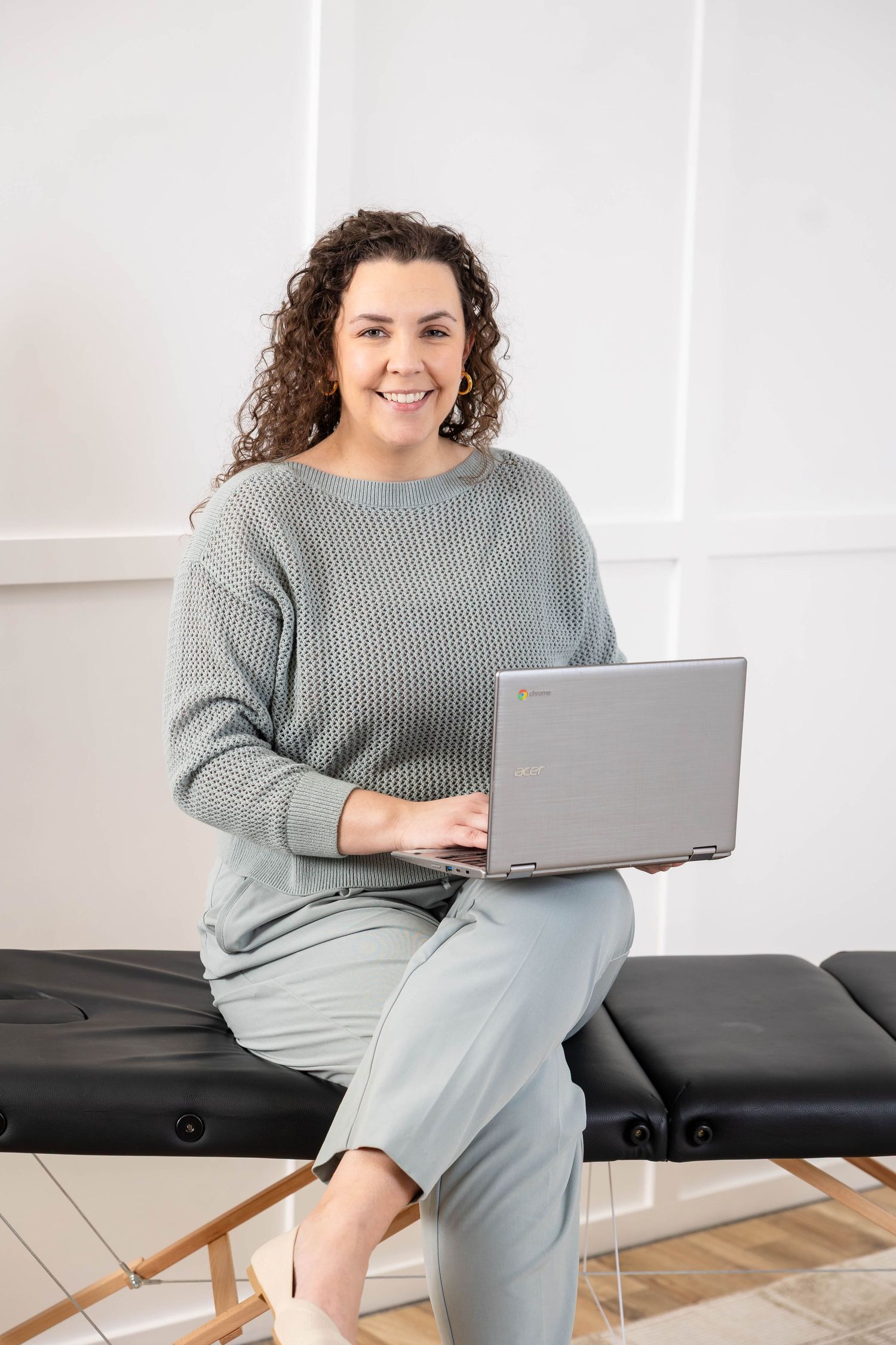Smiling woman with curly hair sits on a black bench holding a laptop, wearing a gray sweater in a bright white room.