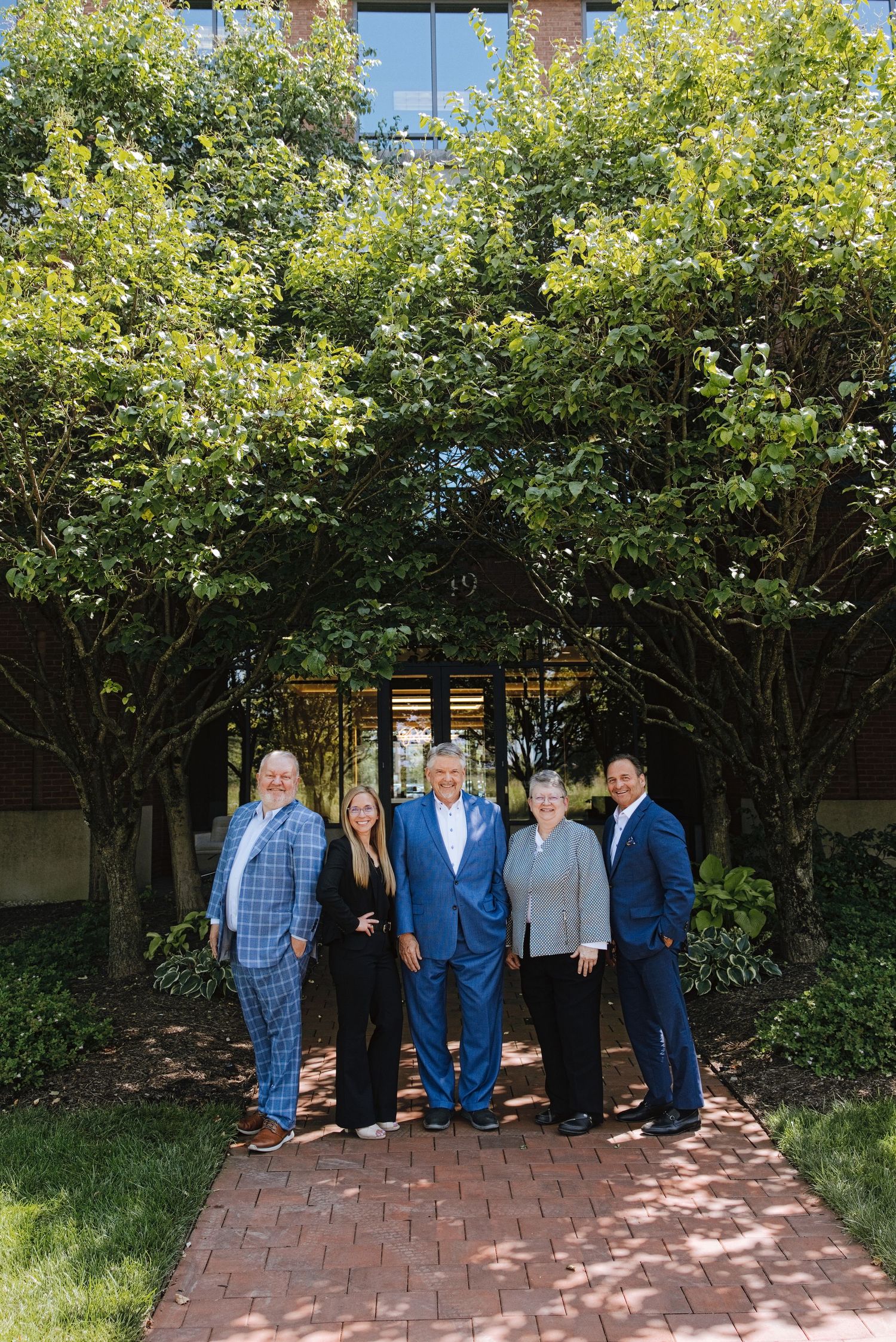 Five professionals posing together on a brick path surrounded by lush green trees outside a modern building.