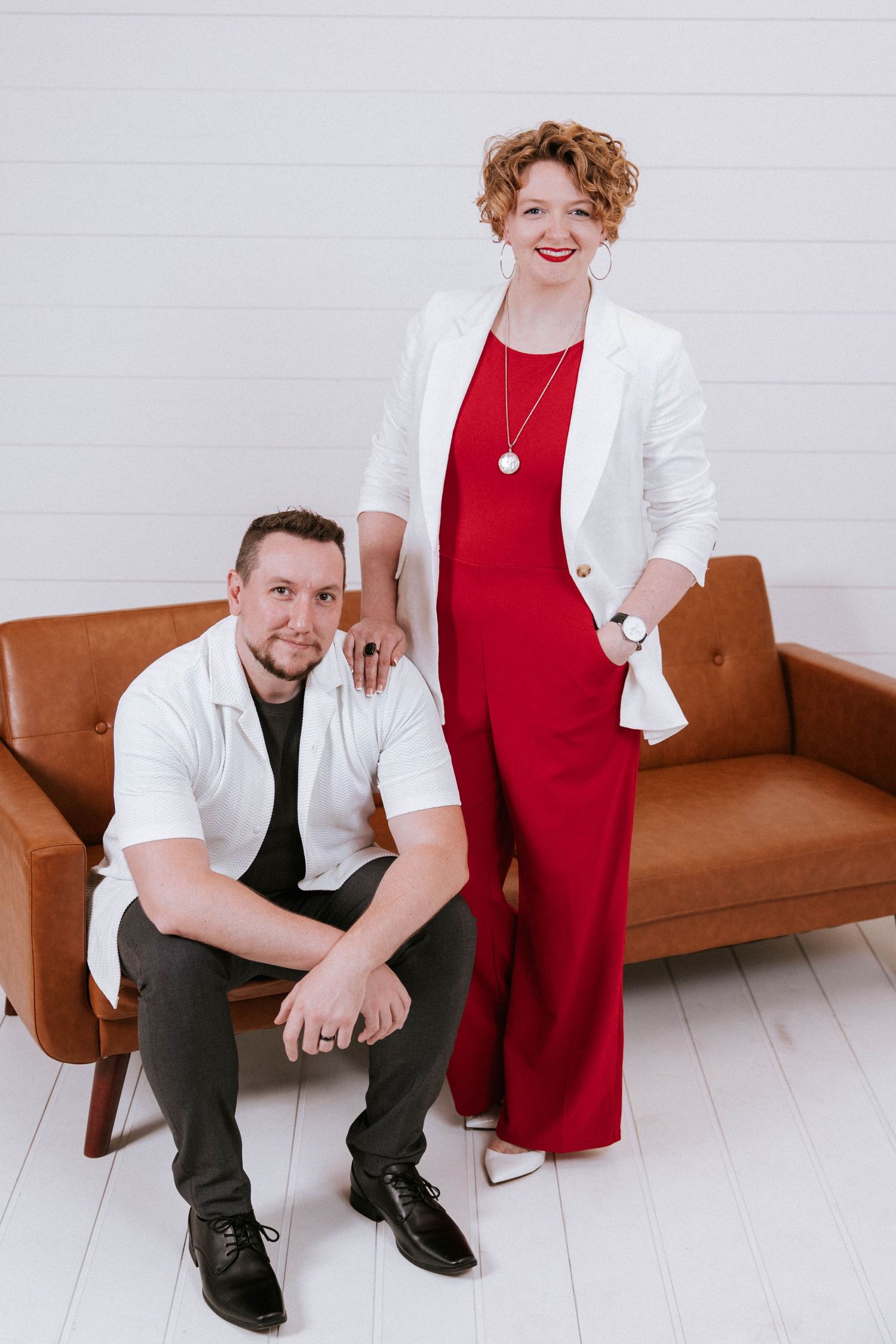 A man in white shirt sits on a leather sofa beside a woman in red jumpsuit and white blazer posing together.