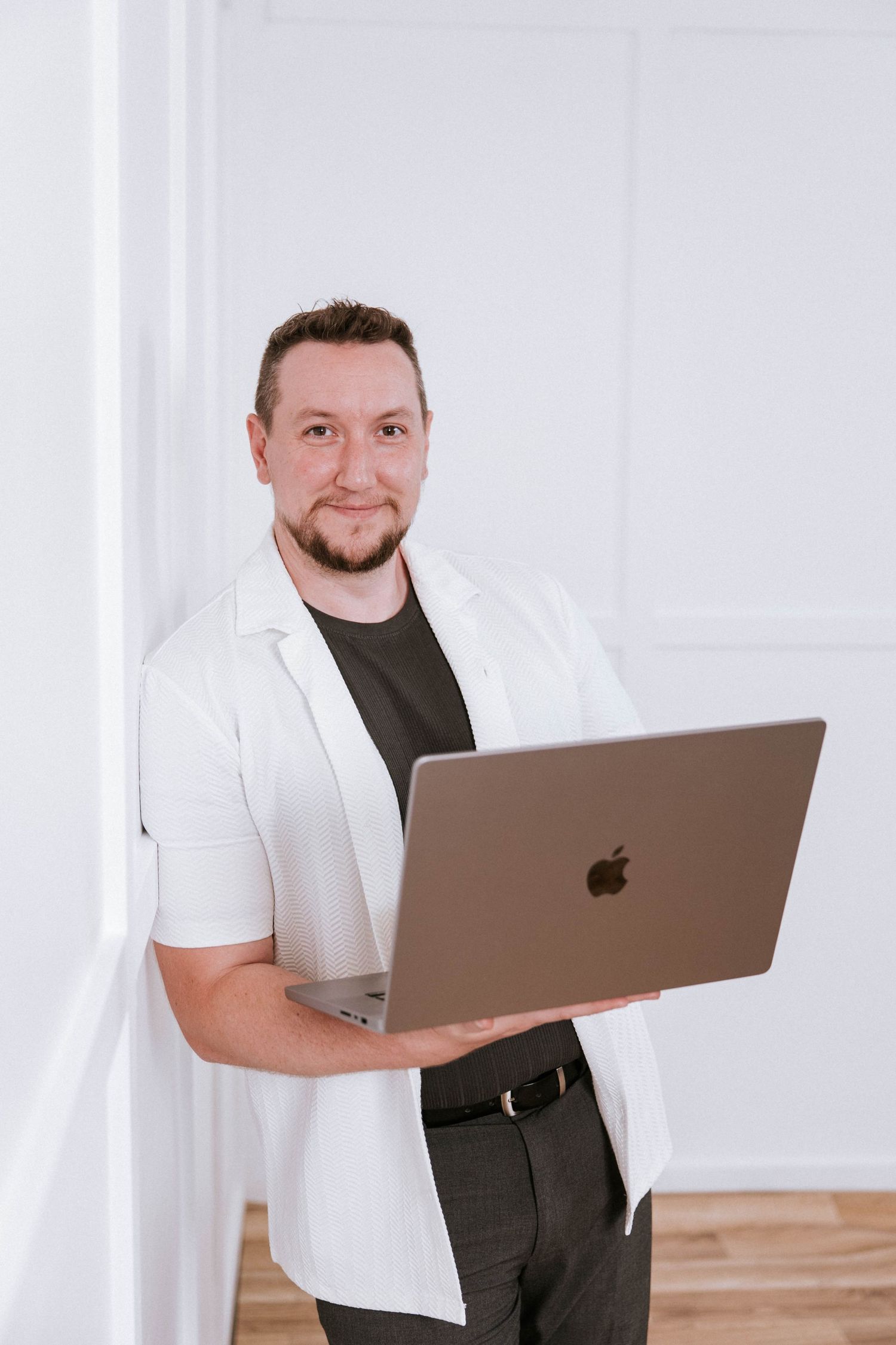 Man in white blazer holding Apple MacBook laptop, smiling confidently in bright white modern room.