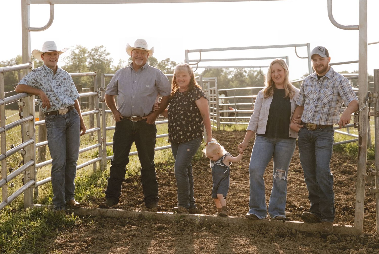 Family of six posing by a cattle gate on a farm, wearing western attire including cowboy hats and jeans.