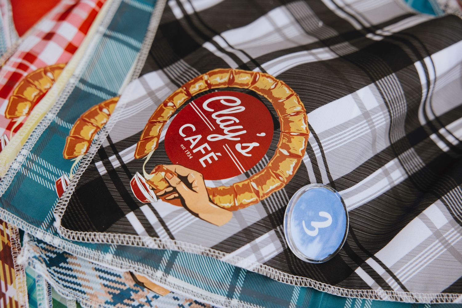 Plaid fabric with a circular cafe logo patch and a small blue button pin on a checkered textile background.