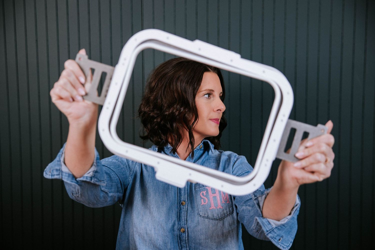 Woman in denim shirt holding a white rectangular frame in front of her face outdoors.