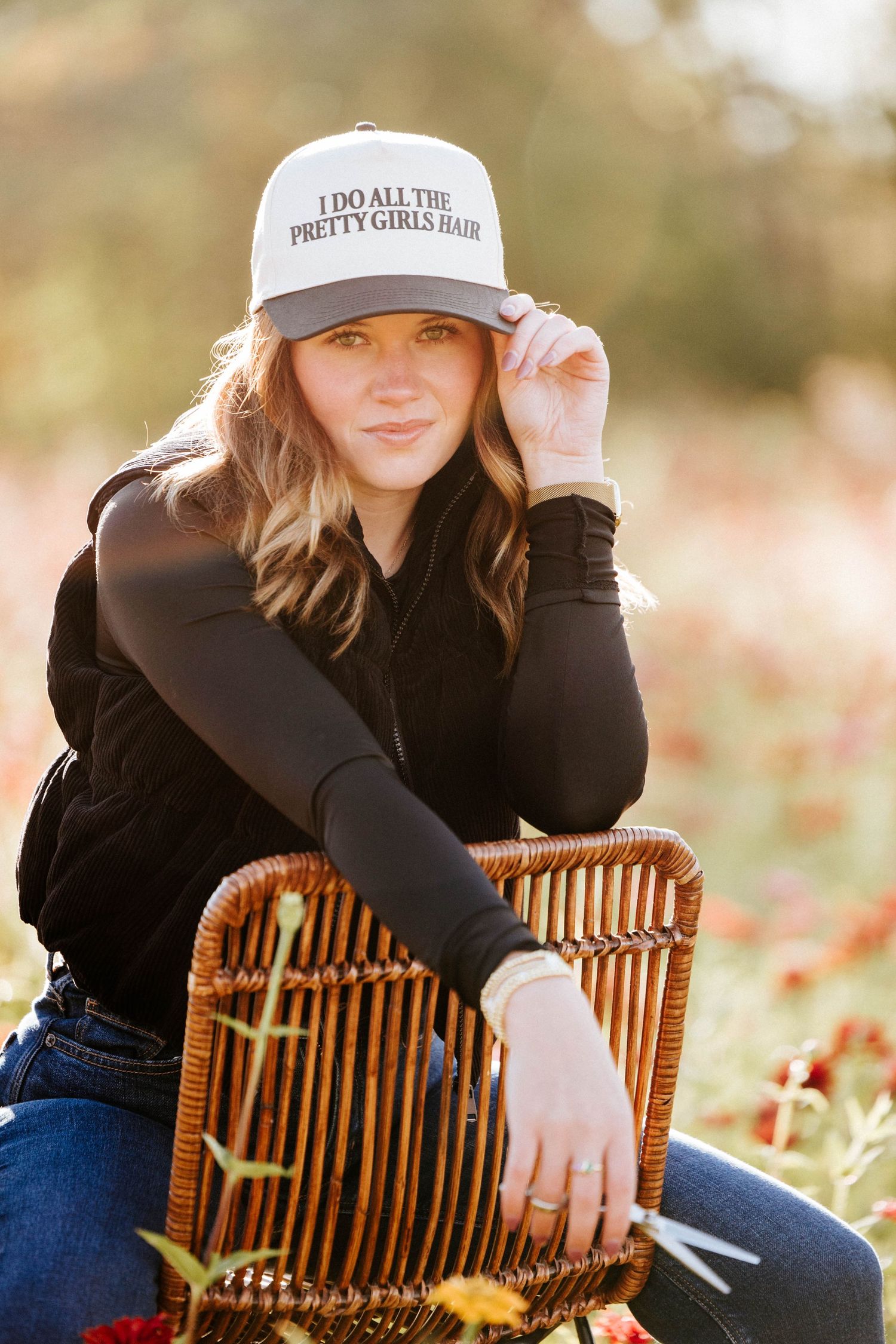 Woman in black outfit and white trucker hat sitting on wicker chair in flower field.
