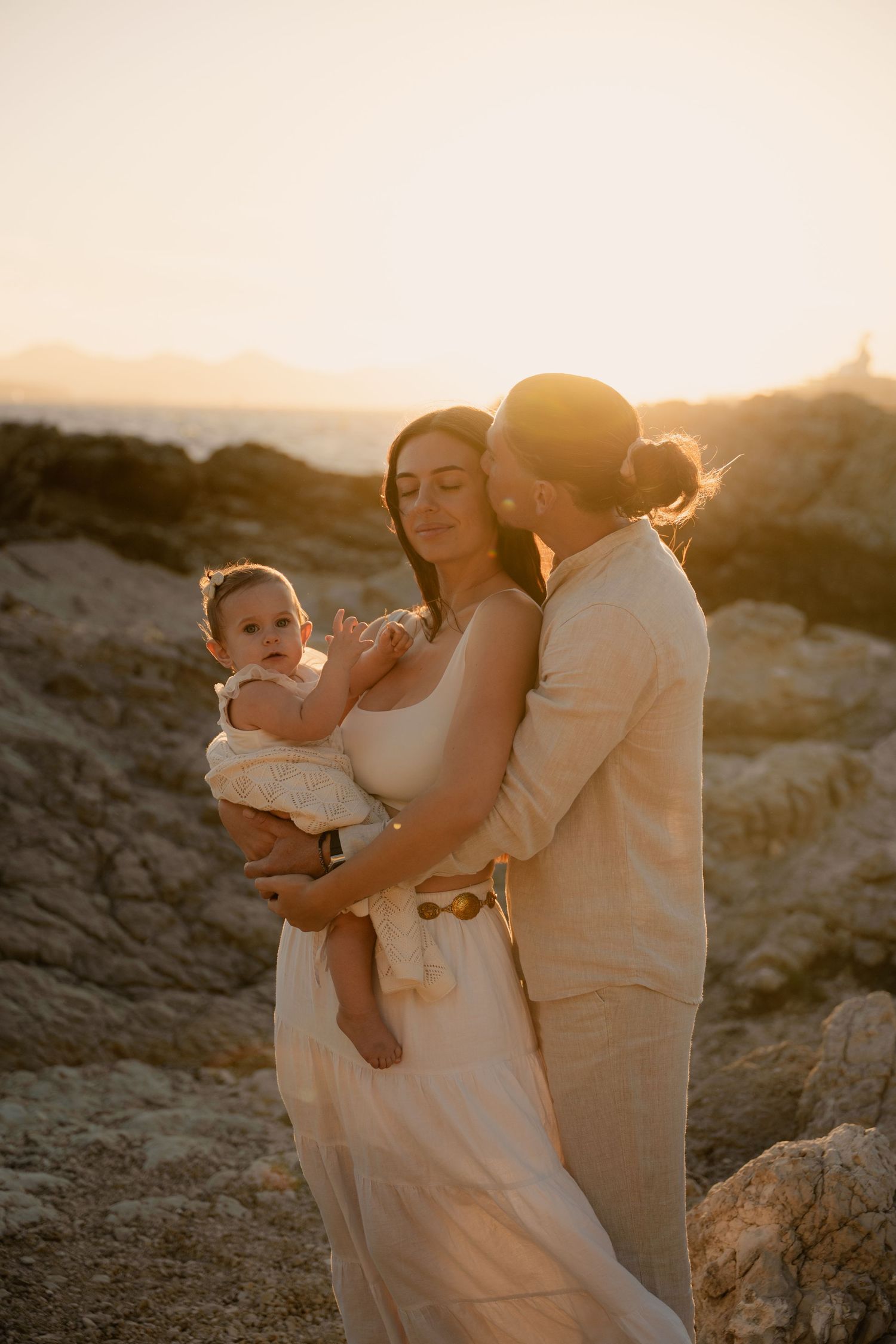 Une famille s'enlace au coucher du soleil, à l'heure dorée, sur un terrain rocailleux, vêtue de vêtements clairs.