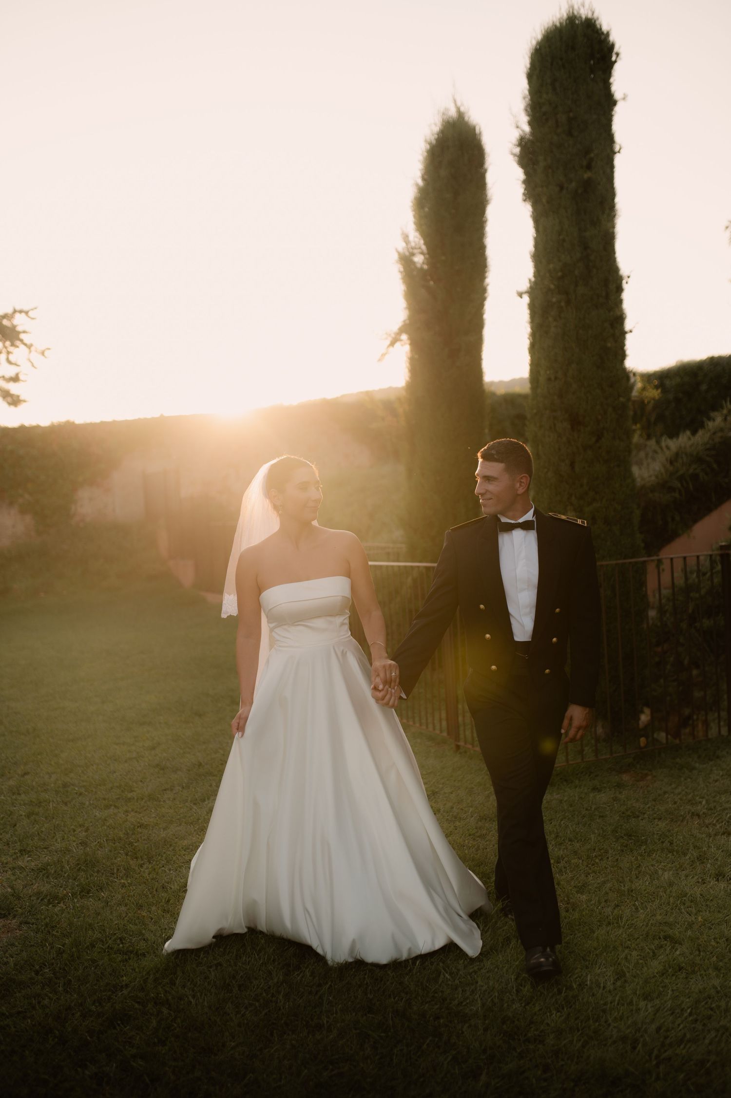 Photo d'un couple de jeune mariés qui marchent en se regardant tendrement  devant les cyprès à la golden hour.