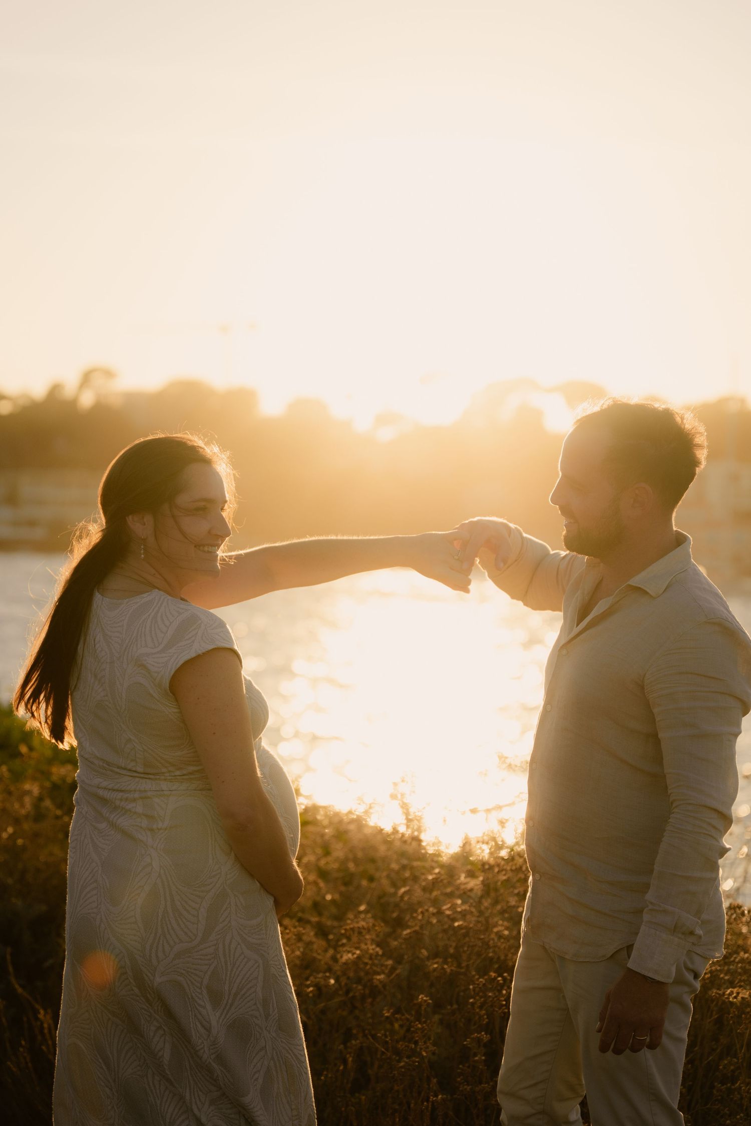 Un couple vêtu de blanc partage un tendre moment au coucher du soleil, au bord de l'eau, on les voit rire en se regardant.