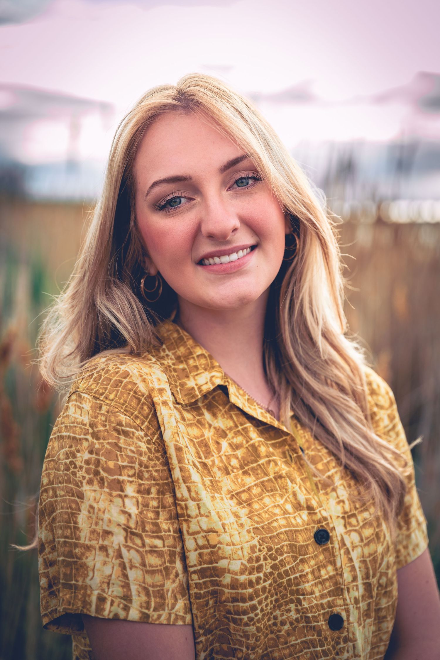 Woman smiling in a field at dusk, wearing a patterned shirt for her graduation portraits.