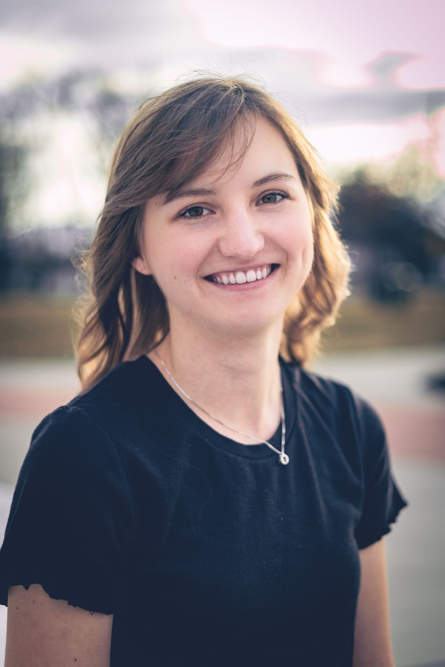 A young woman with shoulder-length hair and a black t-shirt smiles at the camera. She is outdoors with trees and a cloudy sky in the background, creating the perfect setting for dynamic portraits.