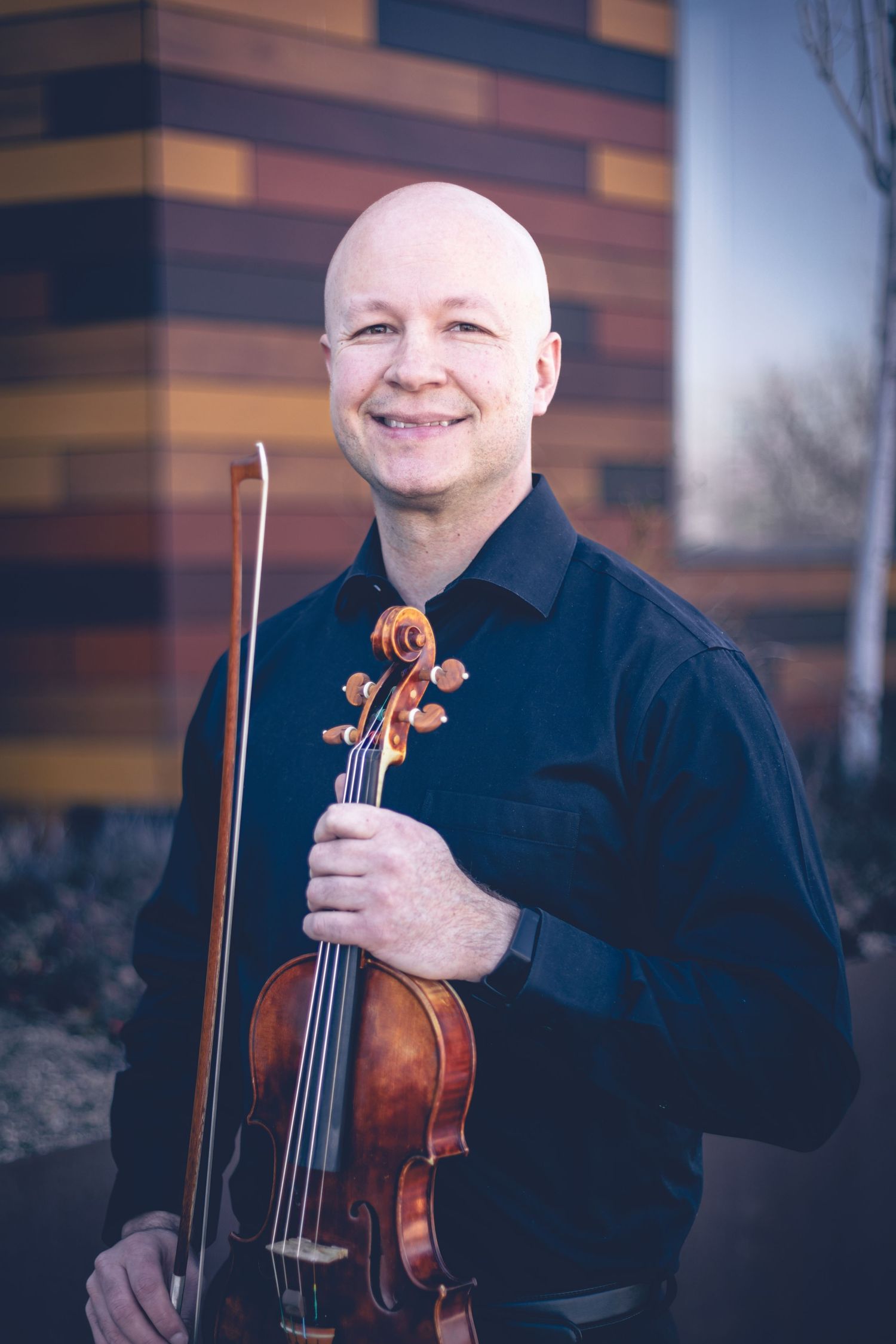 A bald man in a black shirt smiles while holding a violin and bow, standing in front of a building with a multi-colored panel facade, perfect for musician headshots.