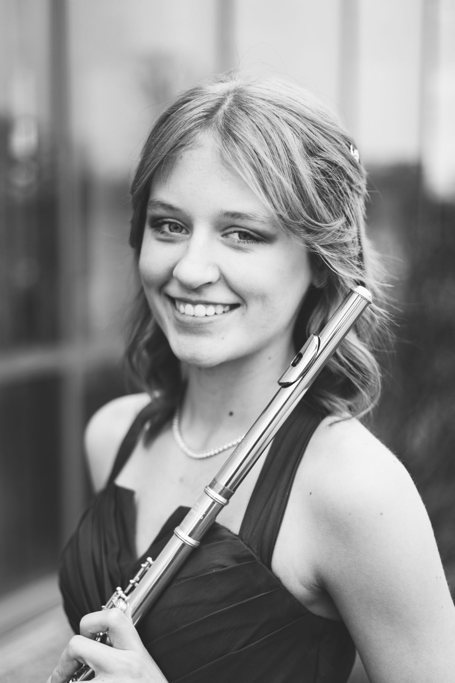 Black-and-white portrait of a smiling person holding a flute, wearing a sleeveless dress and a pearl necklace. They have shoulder-length wavy hair and are standing in front of a blurred background, capturing the essence of musician headshots in dynamic portraits.