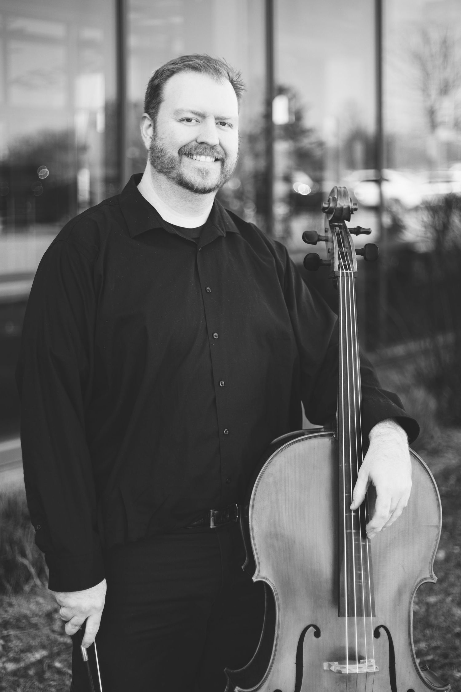 A man with a beard stands outside a building, smiling and holding a cello upright. Dressed in a black shirt and black pants, he exudes confidence, perfect for dynamic portraits that capture his essence as a musician.