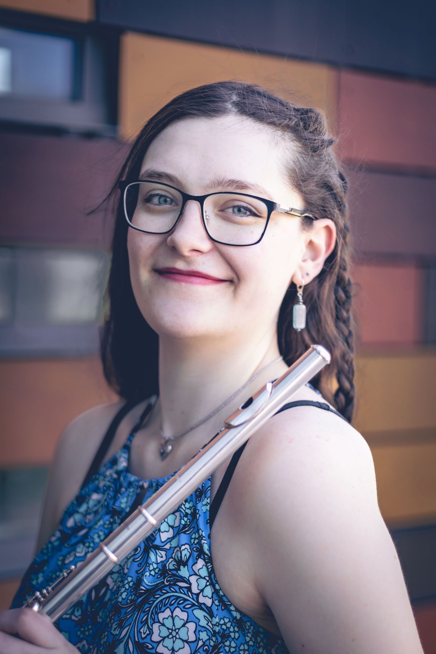 A woman with glasses and a blue patterned top holds a flute while smiling at the camera, perfect for musician headshots. She stands in front of a building with multi-colored panels.