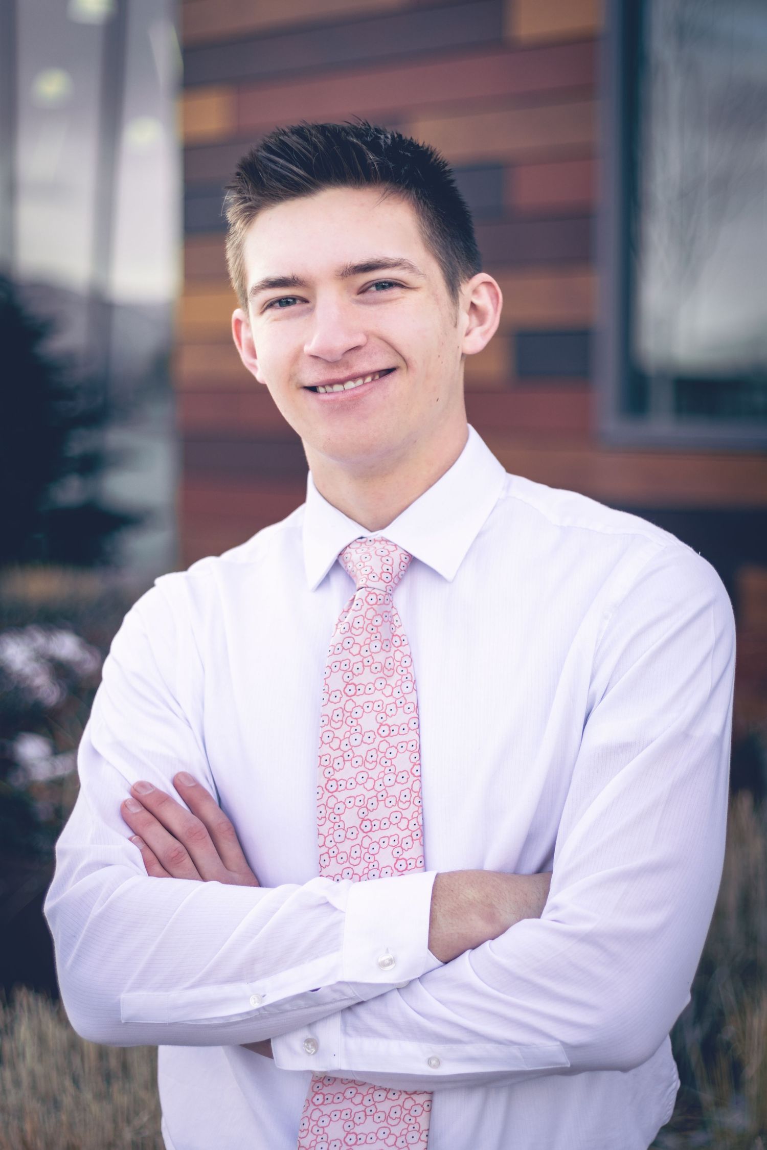 A young man in a white shirt and patterned tie, smiling with his arms crossed, stands outdoors for his senior portraits with Dillon Workman Photography LLC.