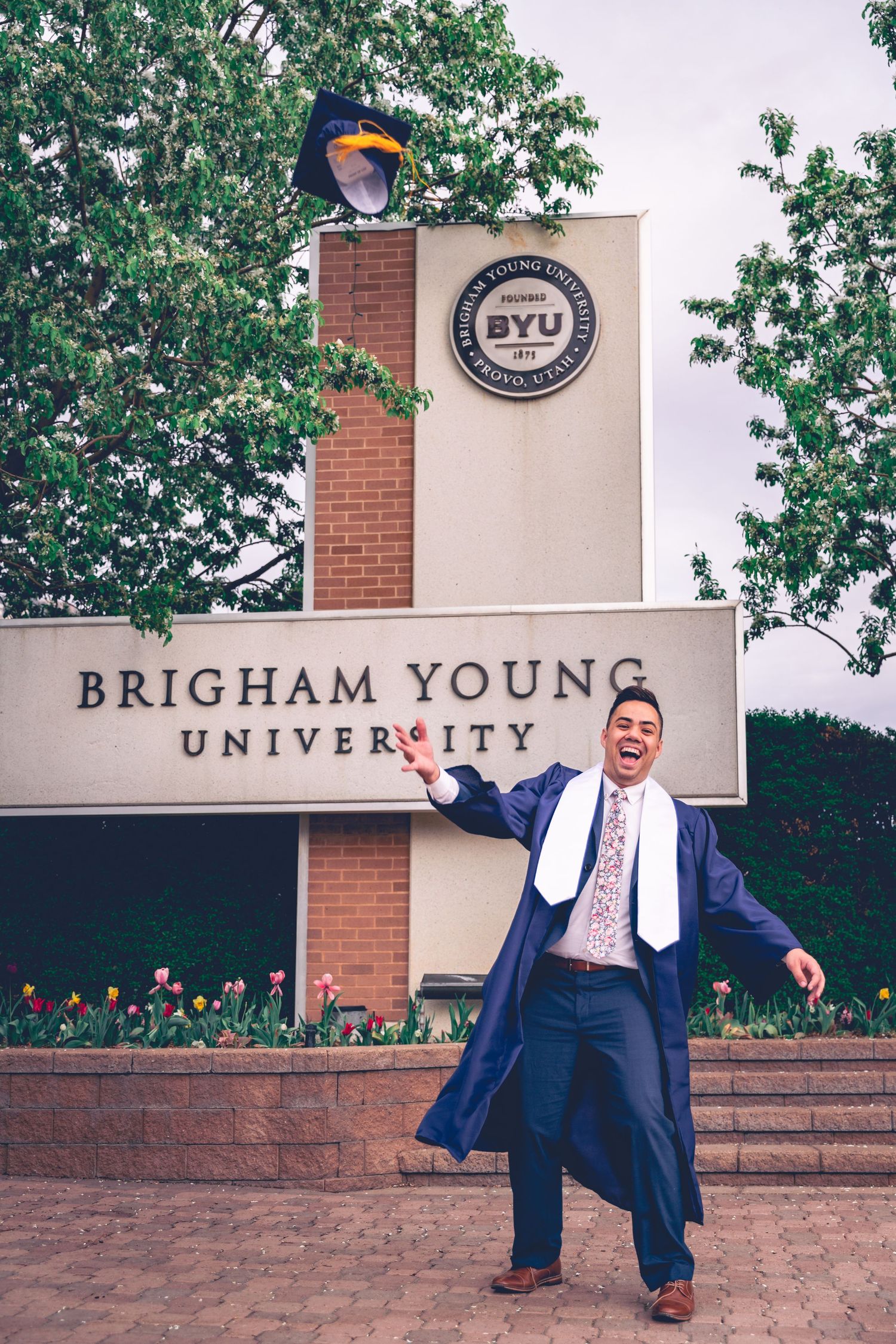 Graduate celebrating in cap and gown in front of Brigham Young University sign for their senior portraits.