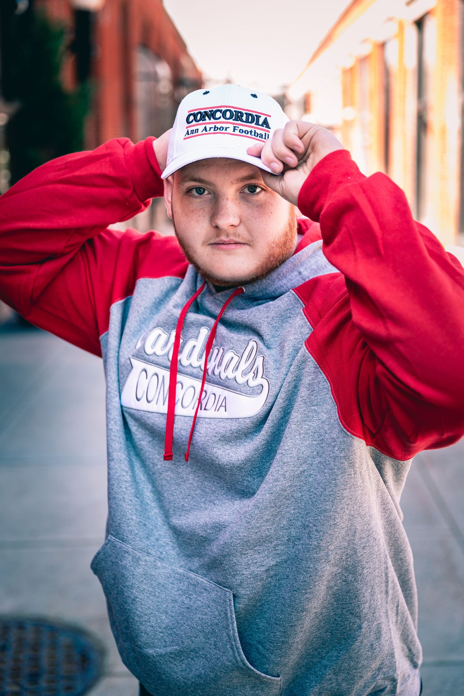 A young man wearing a Concordia University Ann Arbor football cap and hoodie is standing outdoors, looking at the camera with a hand adjusting his cap for his graduation portraits.