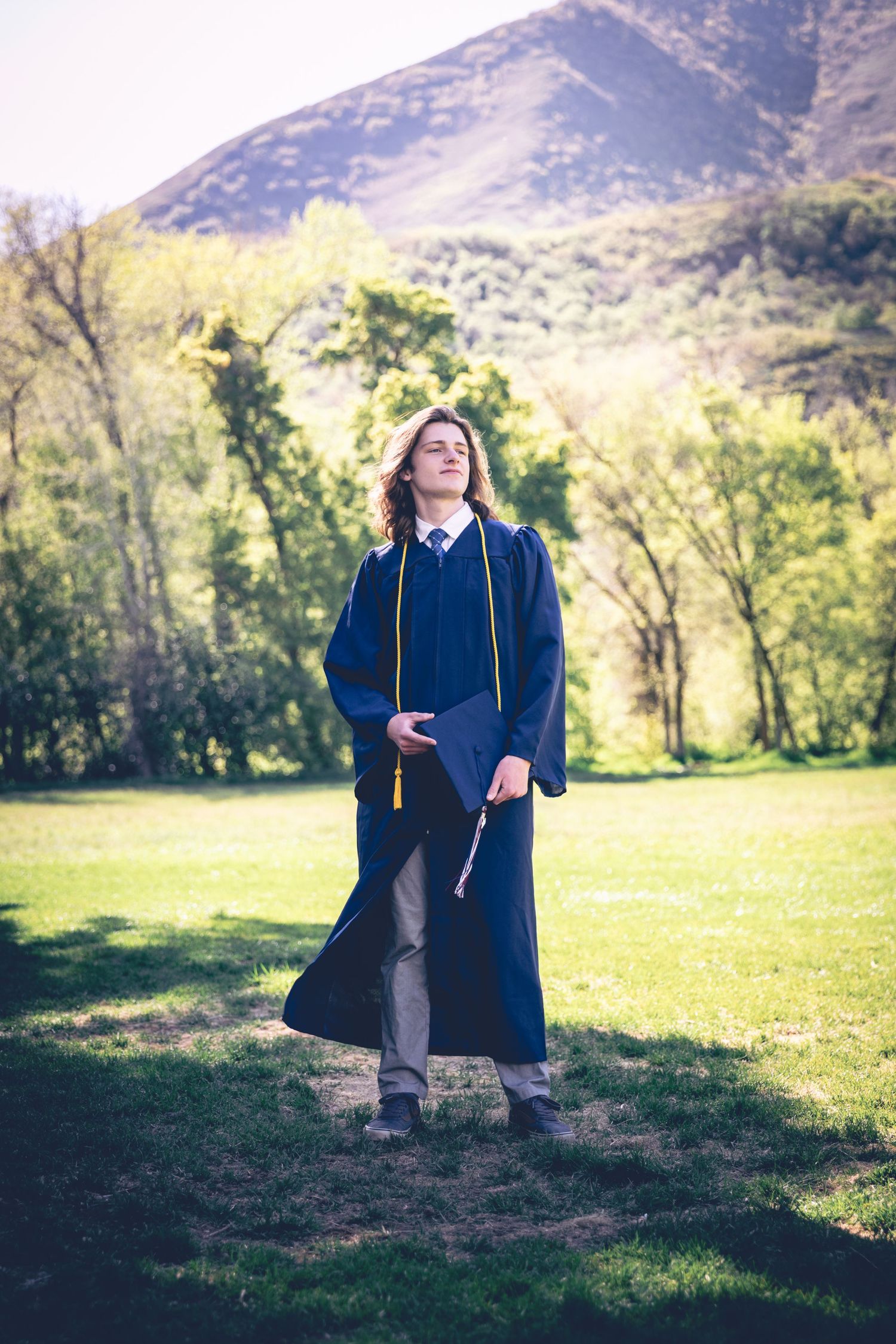 A person in graduation attire stands on a grassy field with trees and mountains in the background, holding a diploma and wearing a cap and gown, perfect for personalized senior portraits.
