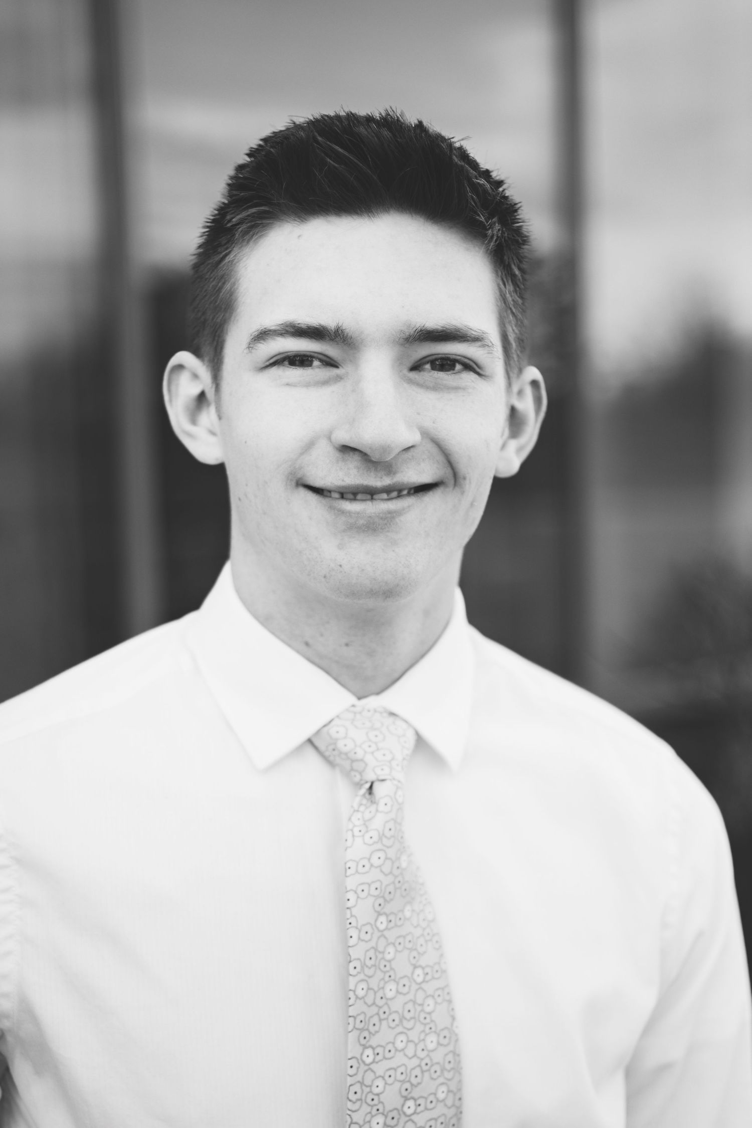 A young man with short dark hair wearing a white dress shirt and patterned tie, smiling in an outdoor setting. The background is blurred, capturing the essence of dynamic portraits.