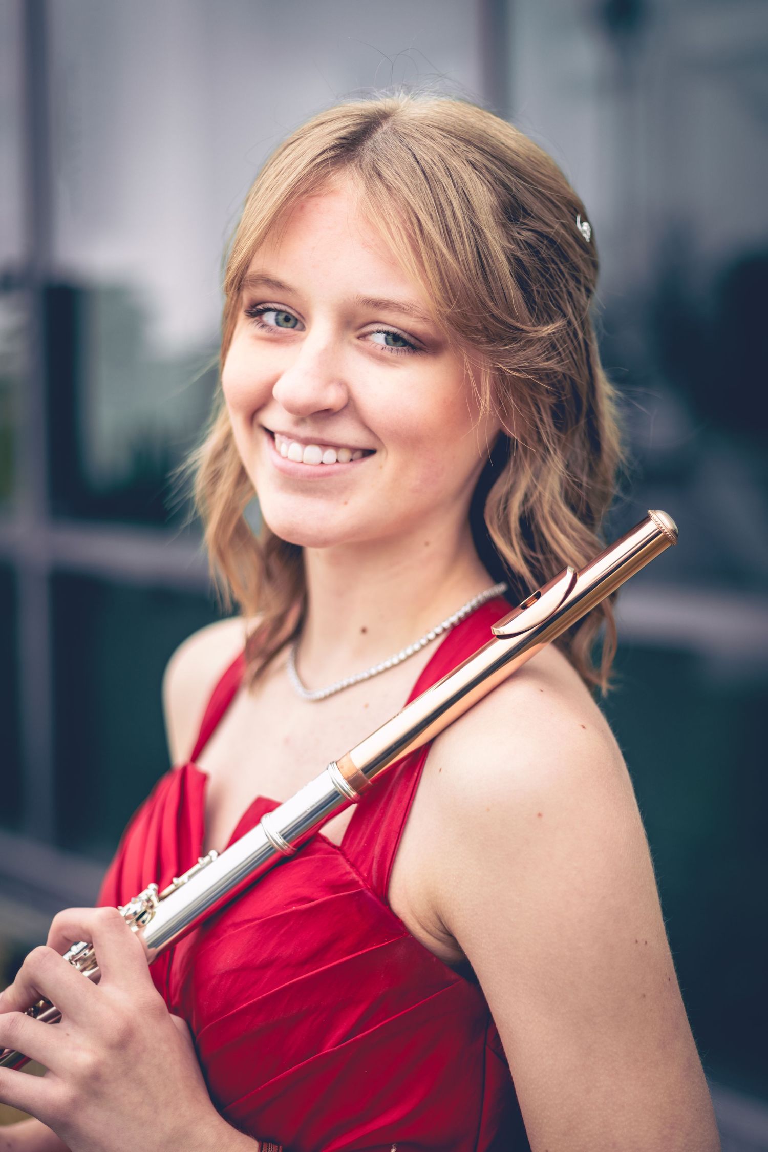 A young woman in a red dress holds a flute and smiles at the camera, standing in front of a building with glass windows, capturing the essence of musician headshots in this dynamic portrait.