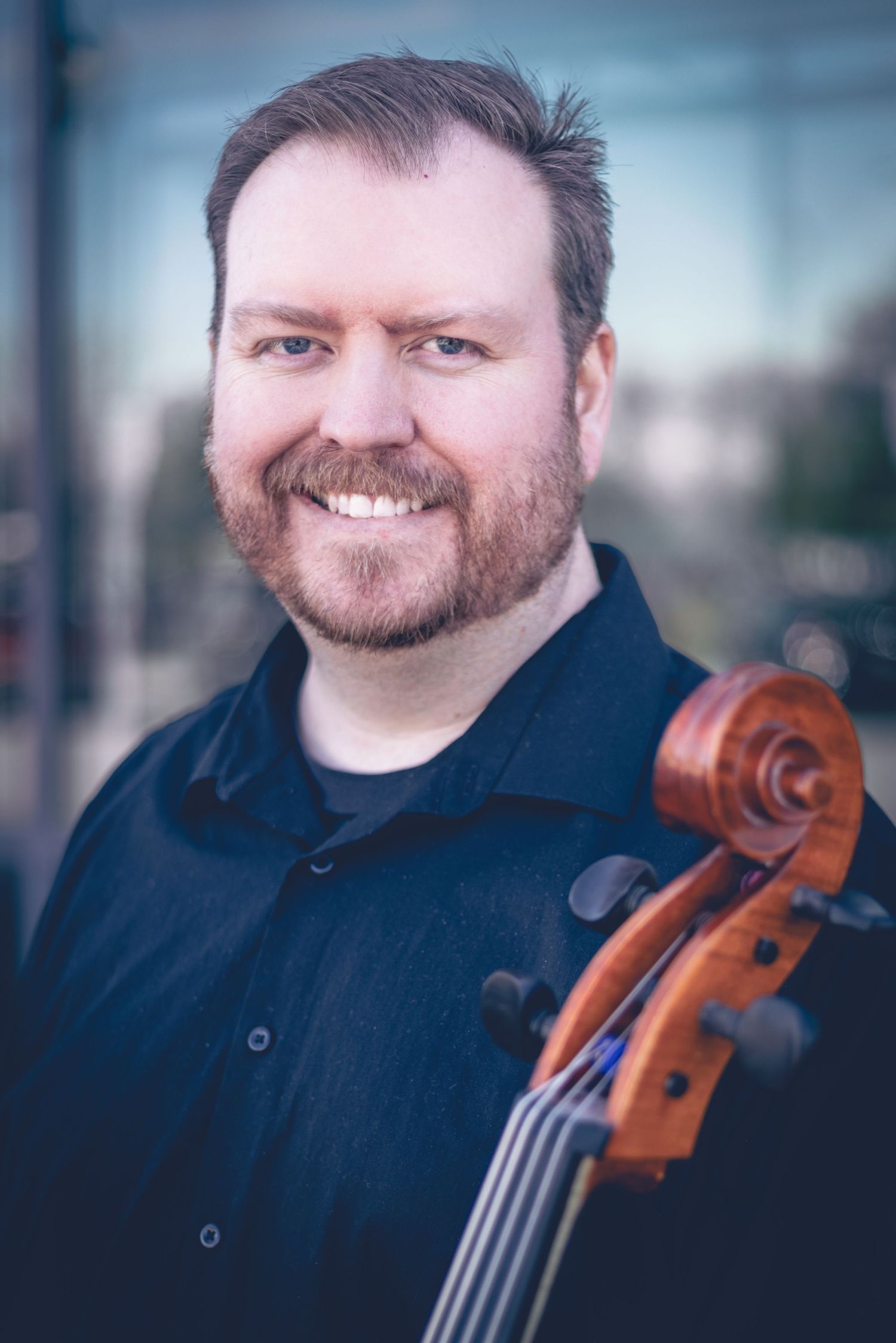 A bearded man smiling warmly, holding a cello, and wearing a black shirt. This musician headshot beautifully captures the essence of personalized sessions, emphasizing both his passion and talent in one dynamic portrait.
