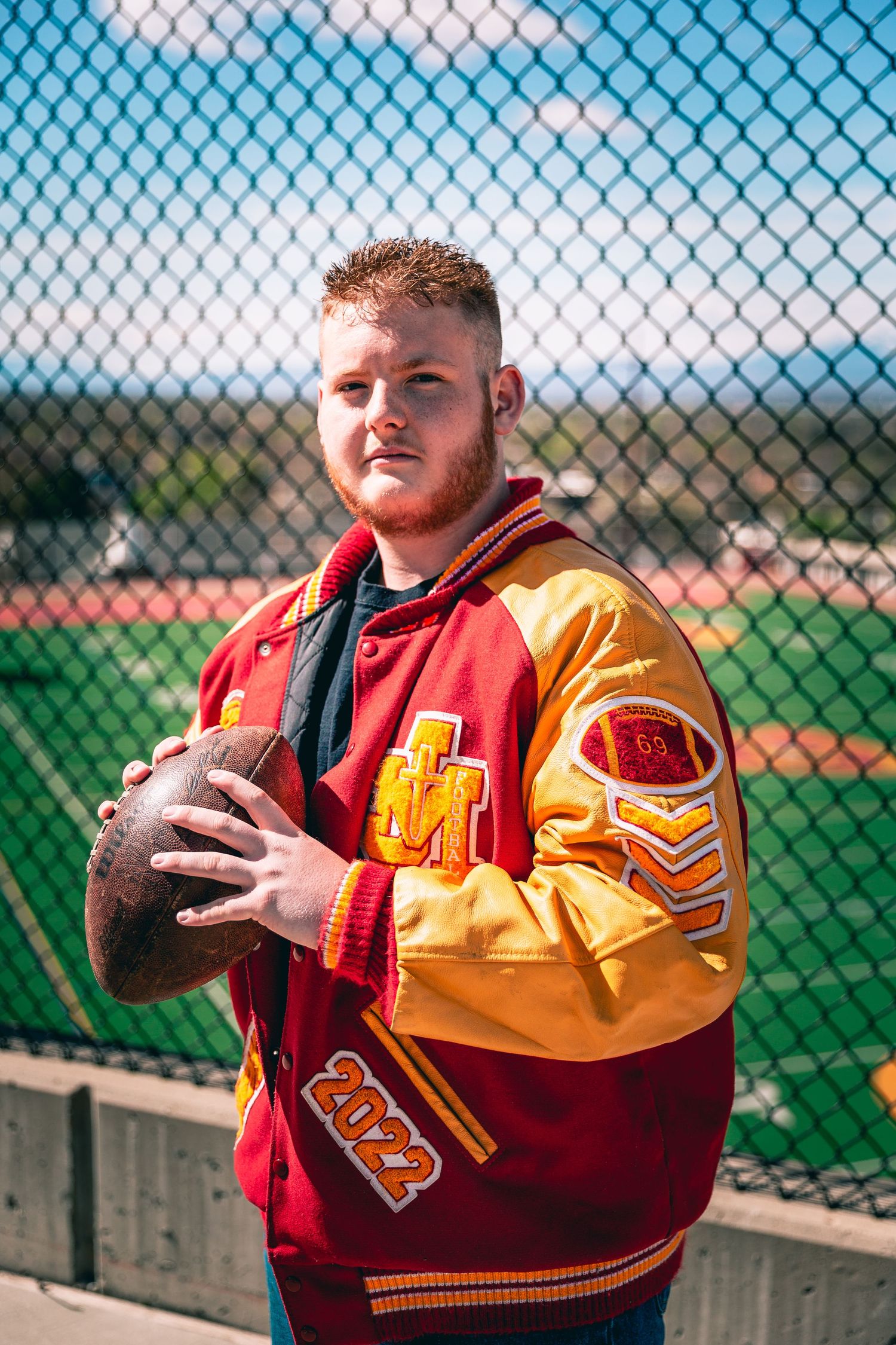Young man holding a football, wearing a letterman jacket by a fence on a sunny day for his personalized senior portraits.