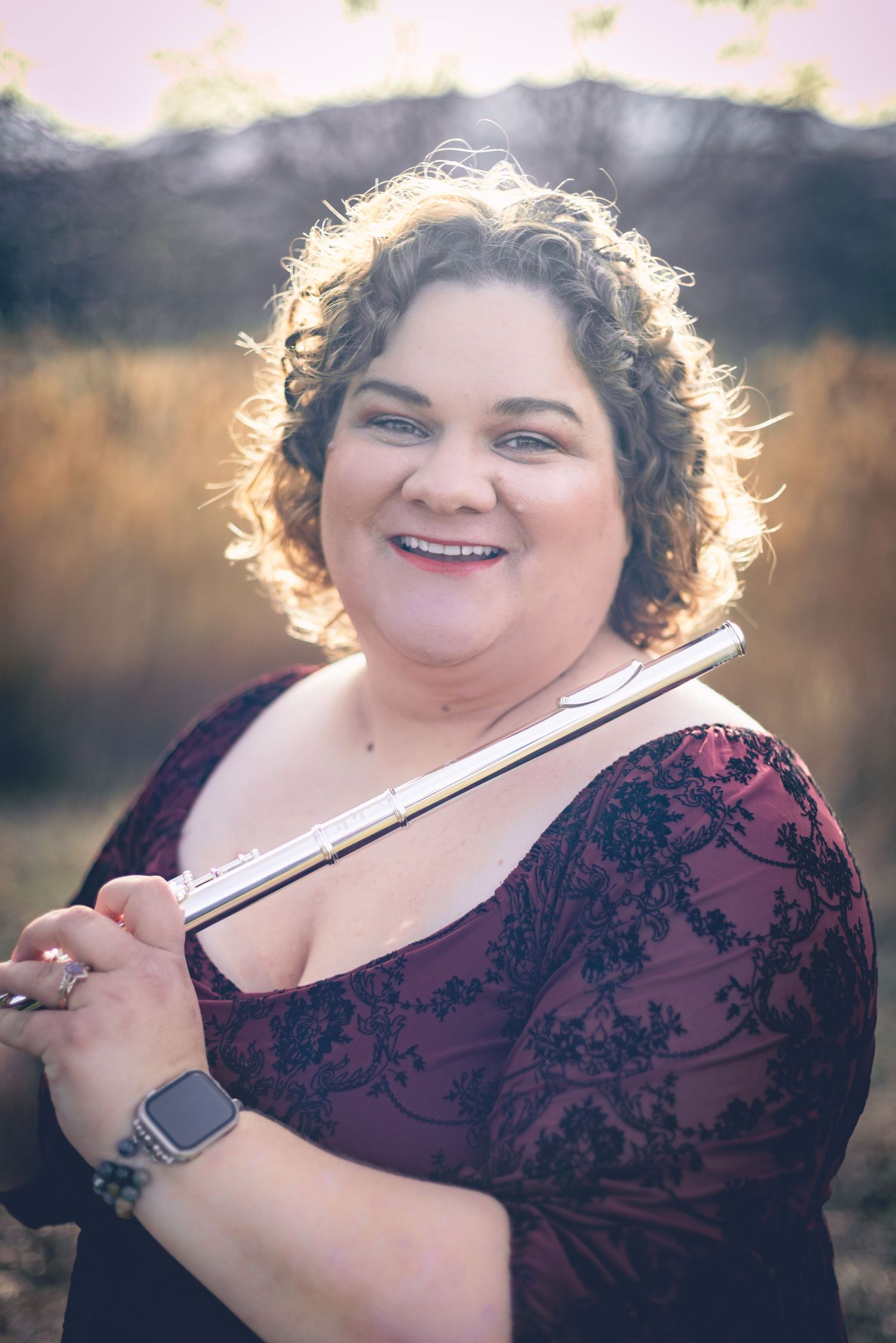 A woman with curly hair holds a flute and smiles at the camera while standing outdoors. She is wearing a maroon dress with a black floral pattern, capturing the perfect blend of style and grace in this musician headshot.