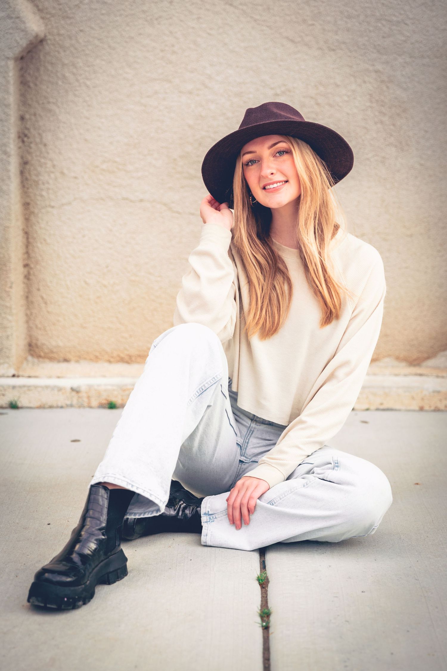 Woman sitting on the ground, smiling, while wearing a hat and casual outfit during her graduation portraits.