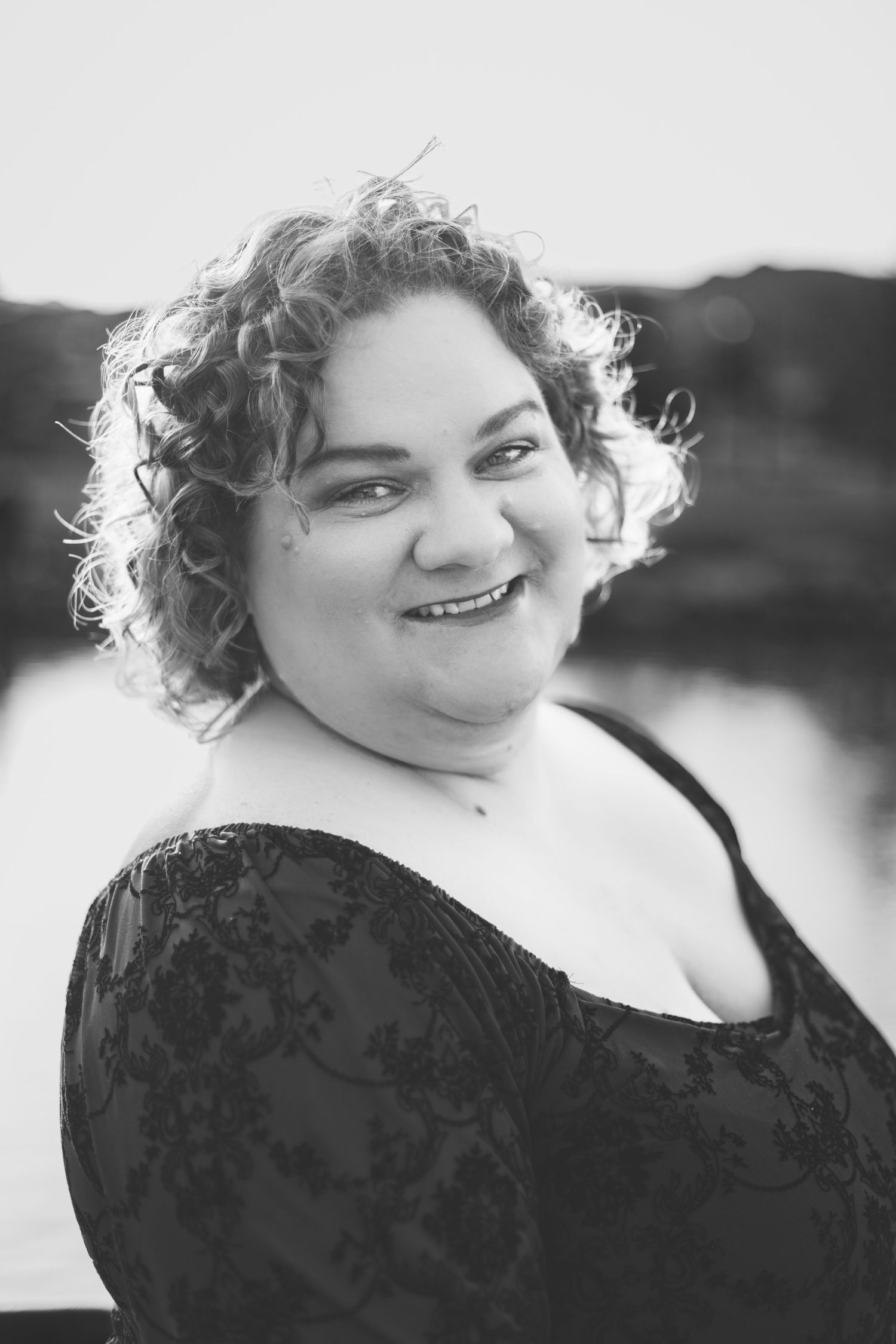 A black and white portrait of a person with curly hair smiling at the camera, wearing an off-the-shoulder top with an intricate pattern. A body of water is visible in the background, creating one of those dynamic portraits that capture both personality and scenery.