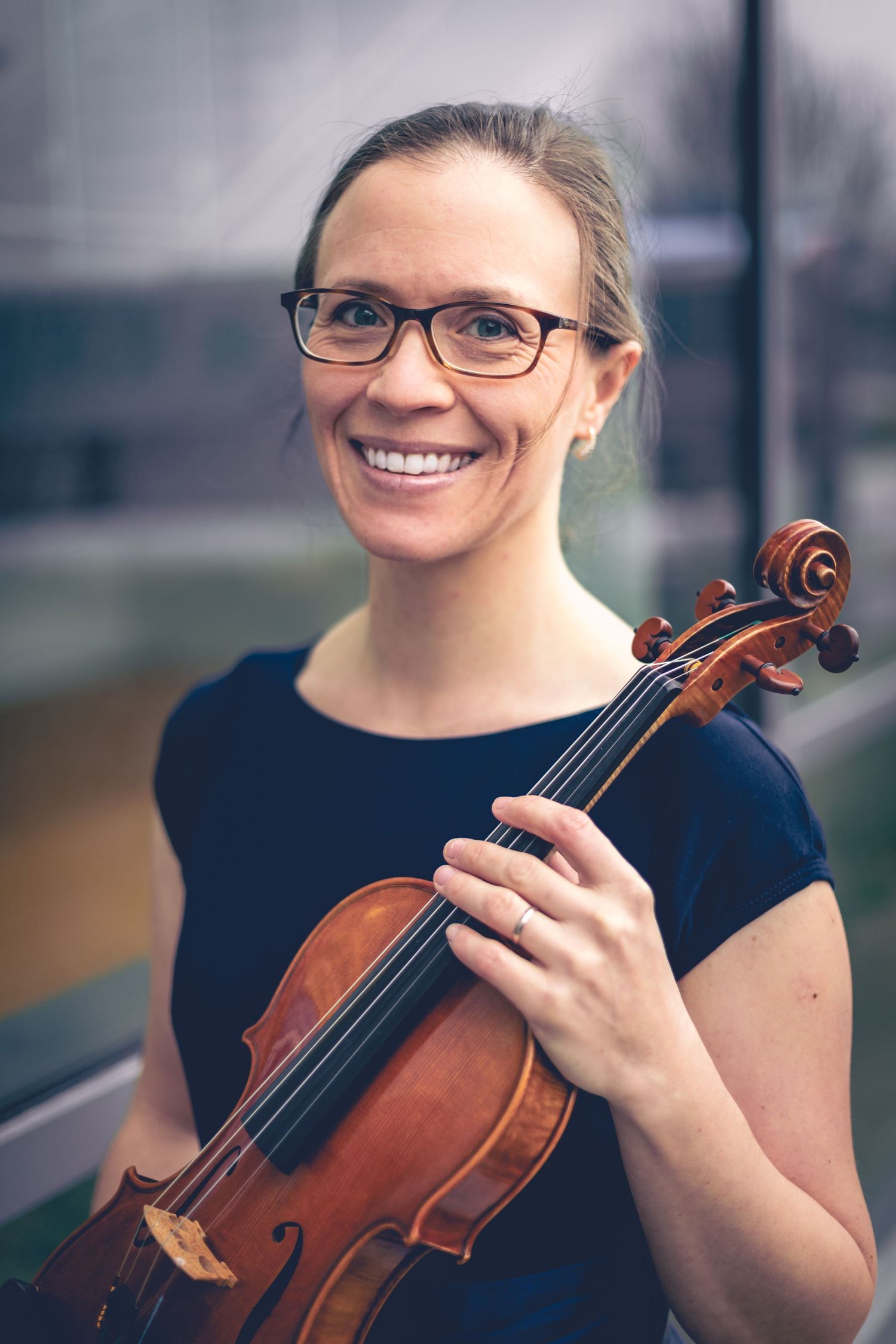 A woman with glasses and a blue dress is holding a violin and smiling, posing for dynamic portraits next to a window.