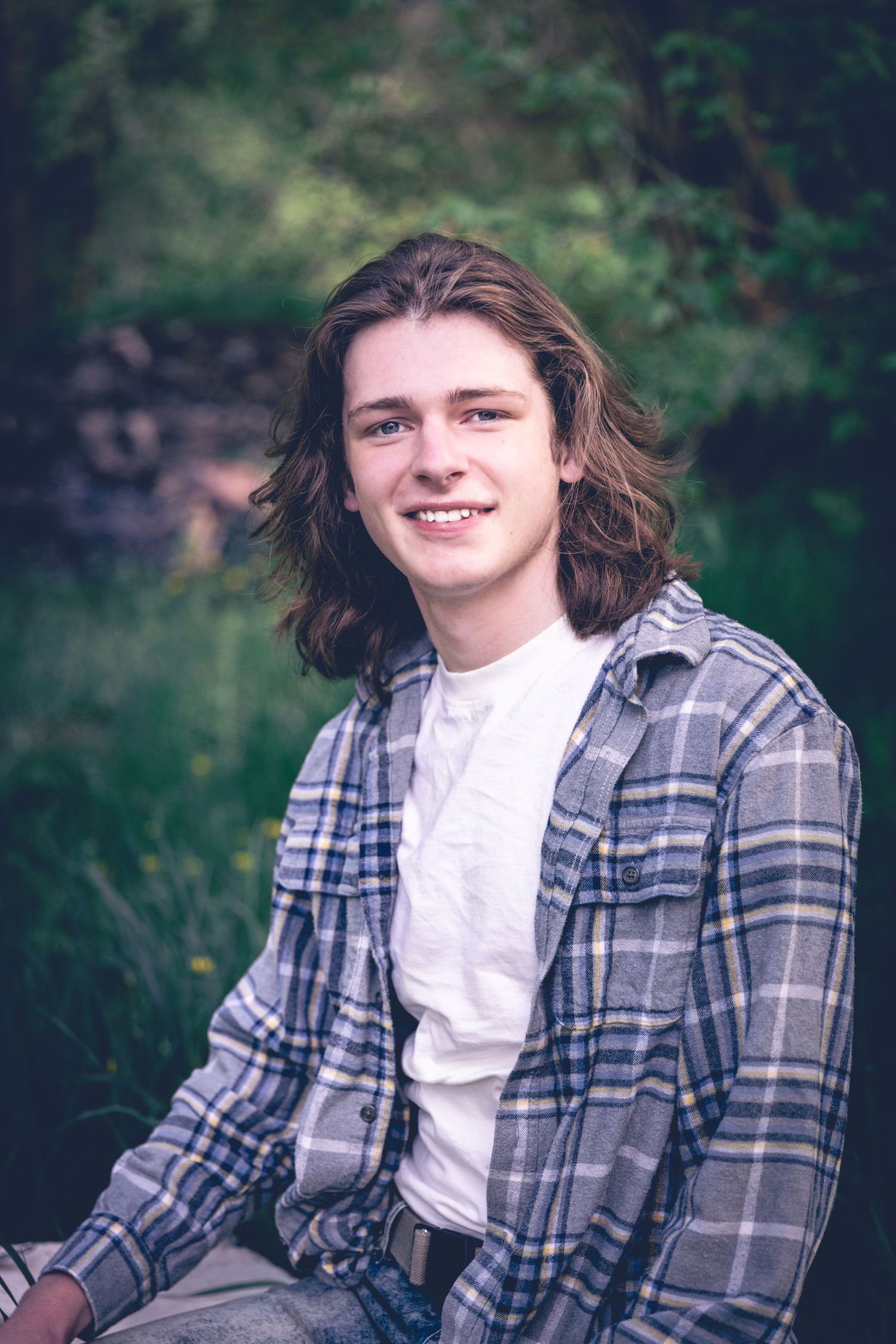 A young person with long hair, wearing a plaid shirt over a white T-shirt, sits outdoors with greenery in the background, capturing the essence of personalized photography perfect for senior portraits.