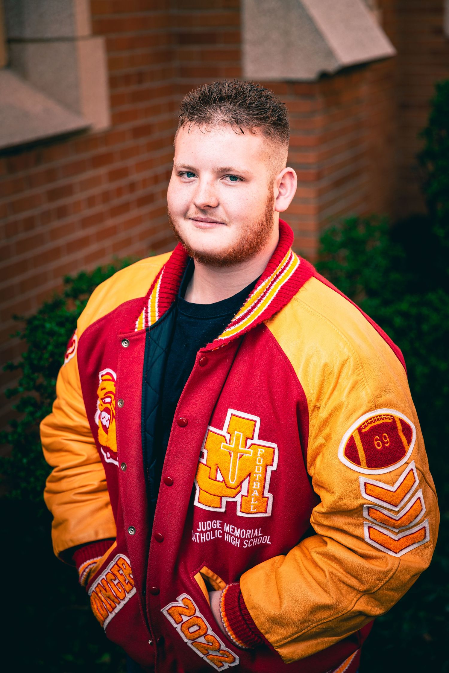 Young man wearing a high school letterman jacket standing in front of a brick wall for his personalized graduation portrait.