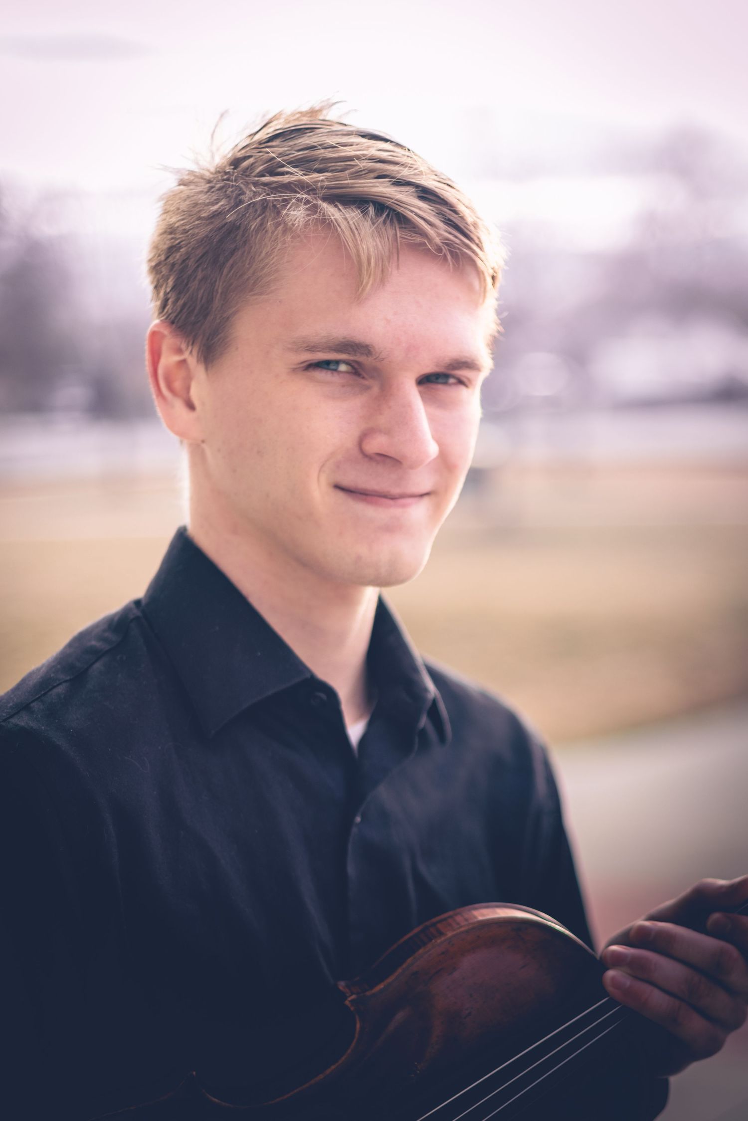 A young man with short blond hair, wearing a black shirt, holds a violin and stands outdoors with a blurred natural background, making for an excellent musician headshot.