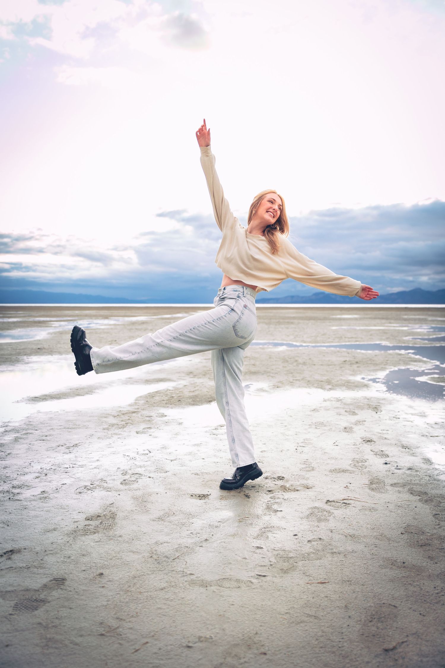 A joyful woman posing playfully on a beach with one arm raised and one leg lifted against a cloudy sky backdrop, in a personalized photography session.
