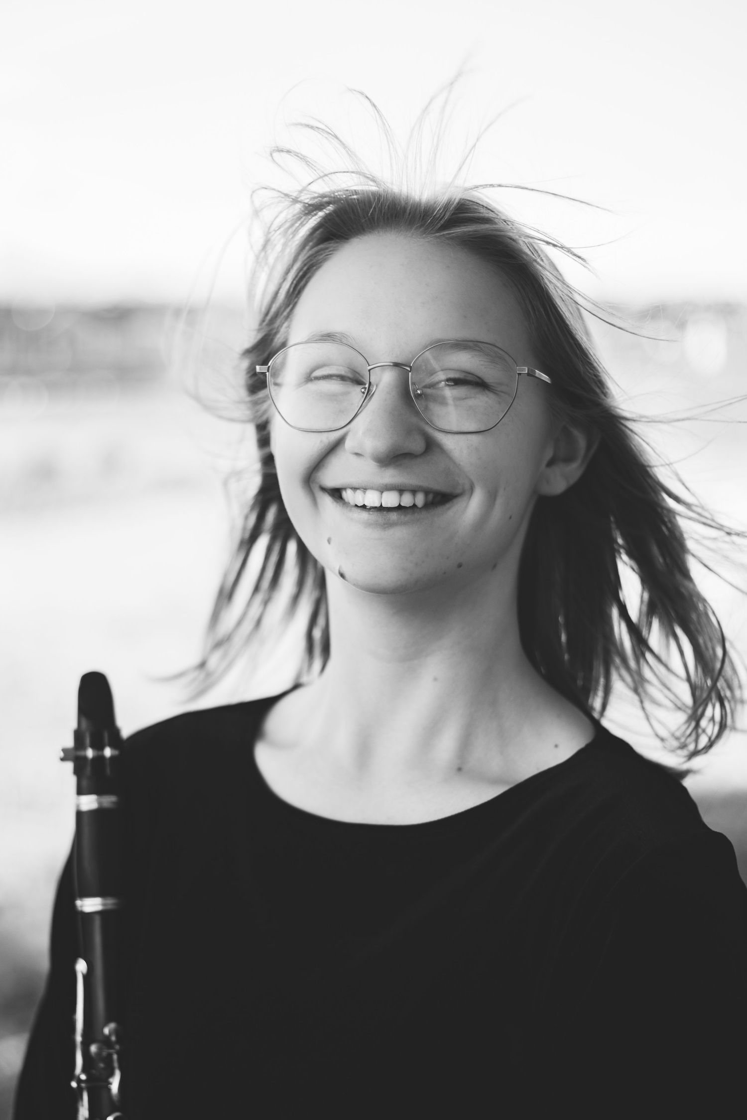 Black-and-white photo of a smiling person with glasses, holding a clarinet. The wind is blowing their hair as they wear a dark top, perfectly capturing the essence of dynamic portraits often seen in personalized sessions for musician headshots.