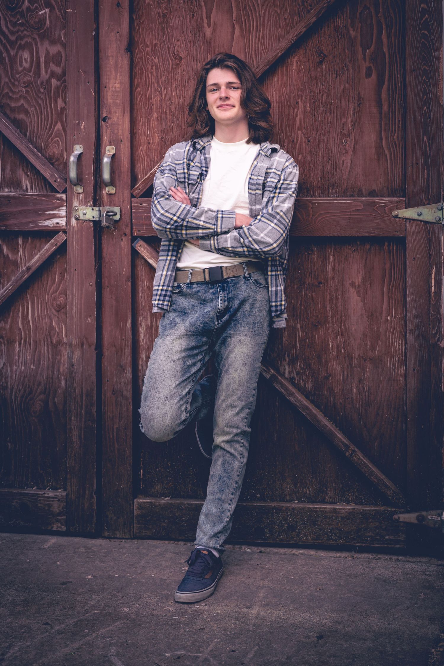 A person with long hair leans back against a wooden door with crossed arms, wearing a flannel shirt, white t-shirt, jeans, and sneakers. This stylish look is perfect for personalized photography and capturing unforgettable senior portraits.