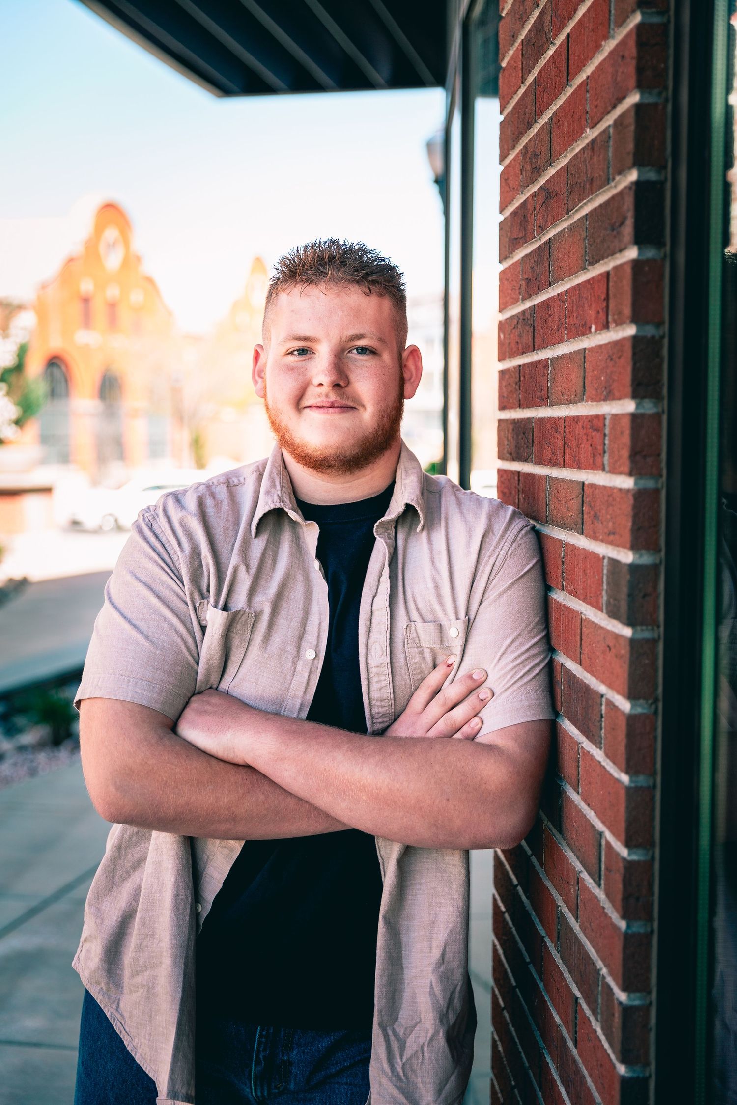 A young man with crossed arms leaning against a brick wall outdoors, showcasing personalized senior portraits