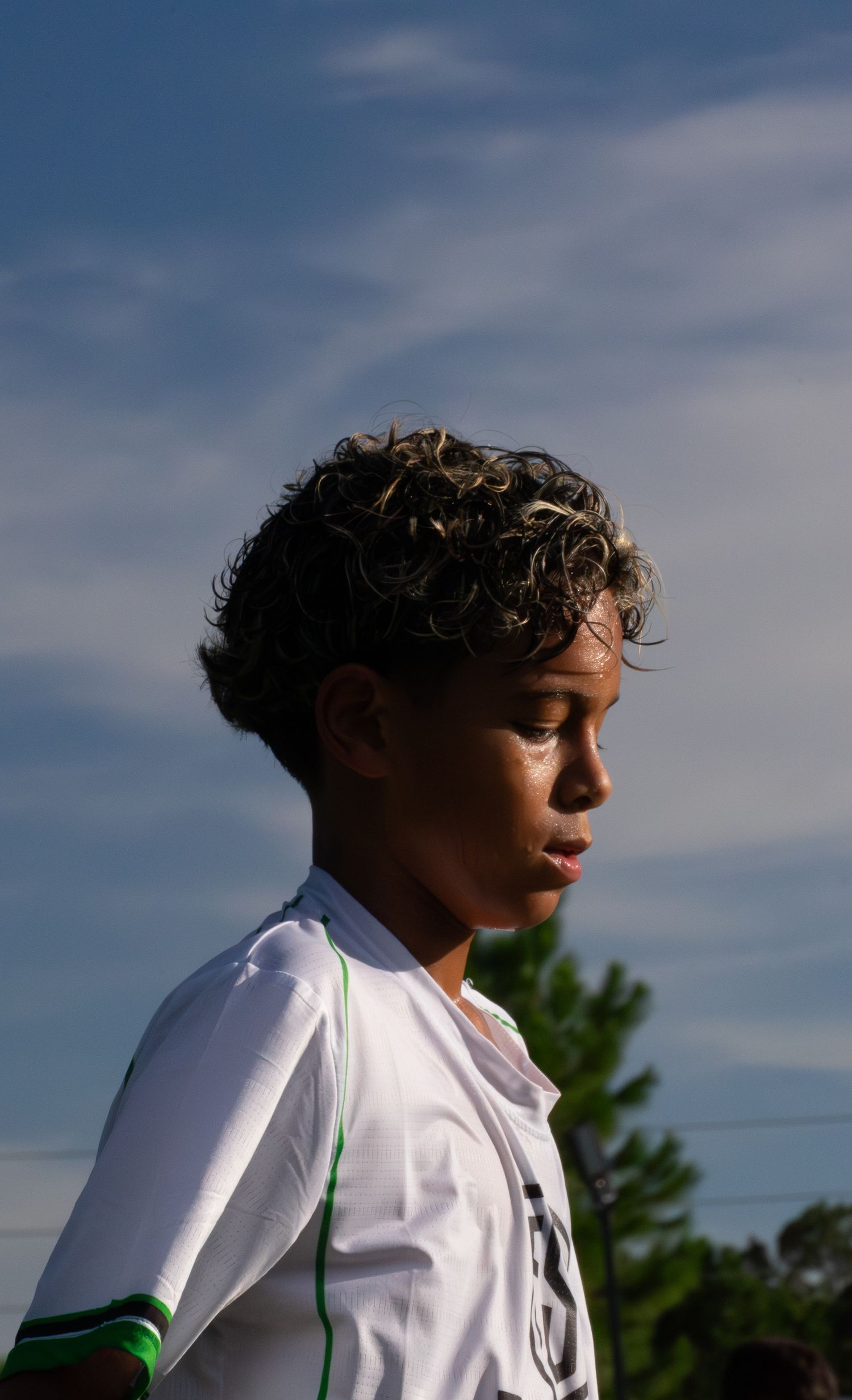 Young curly-haired soccer player in white and green jersey stands against a blue sky backdrop.