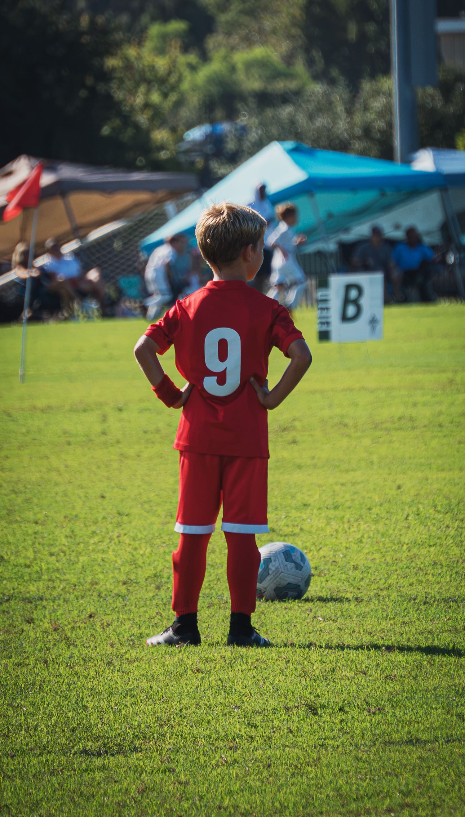 Young boy in red number 9 soccer uniform stands with hands on hips on green field.