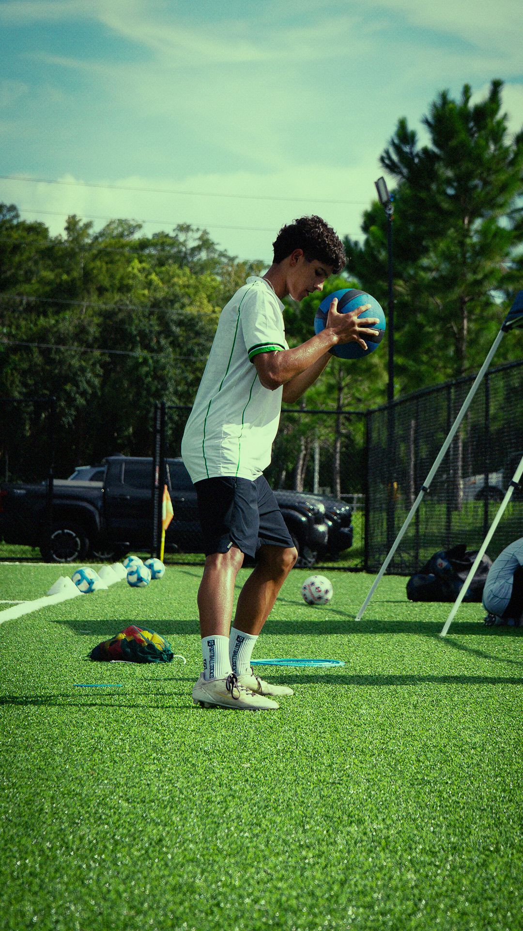 Young soccer player in white jersey holds a medicine ball during training on an artificial turf field.