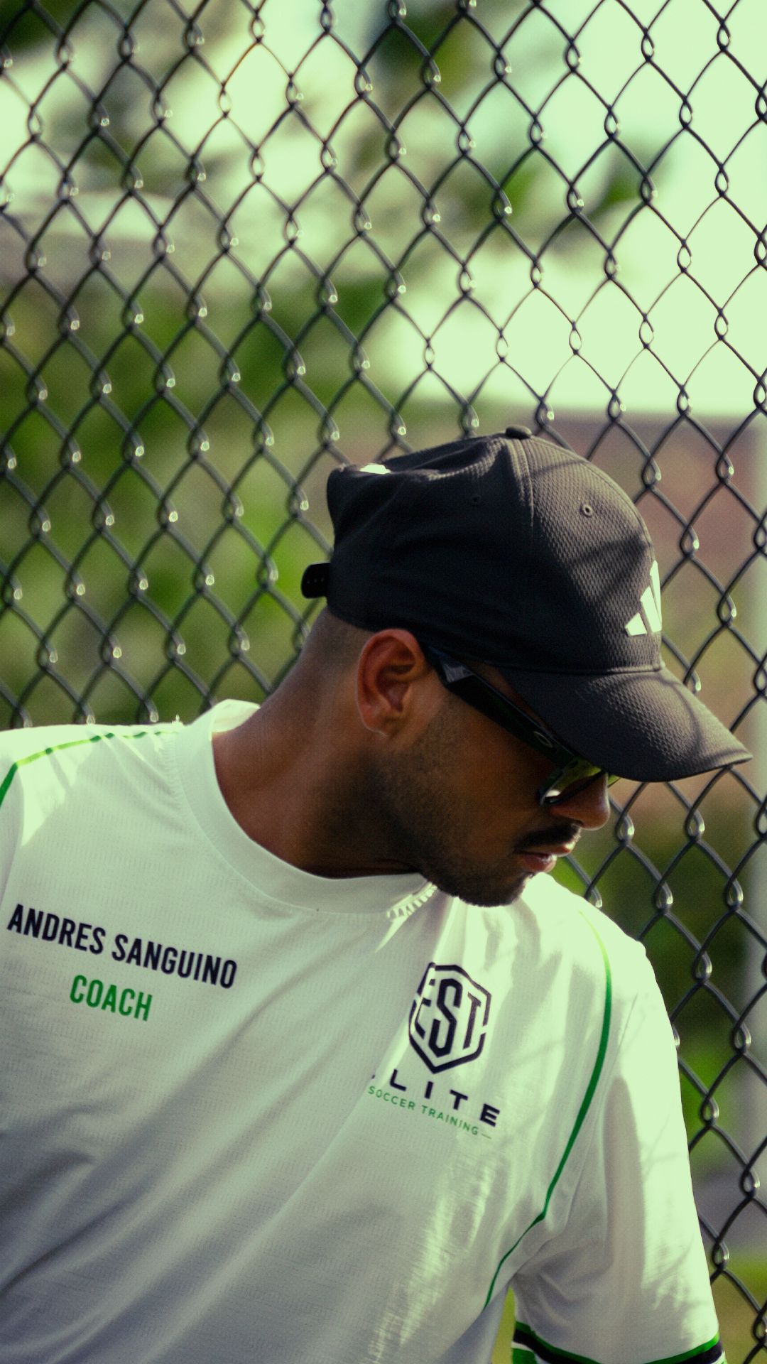 Male soccer coach Andres Sanguino in white Elite Soccer Training shirt stands by chain fence.