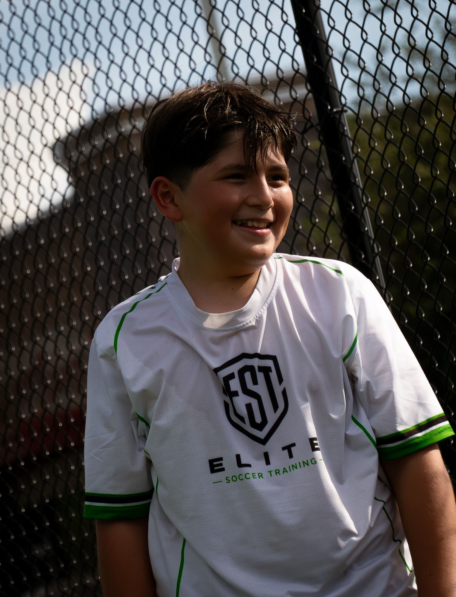 Smiling youth soccer player wearing white Elite Soccer Training jersey stands near a chain-link fence.
